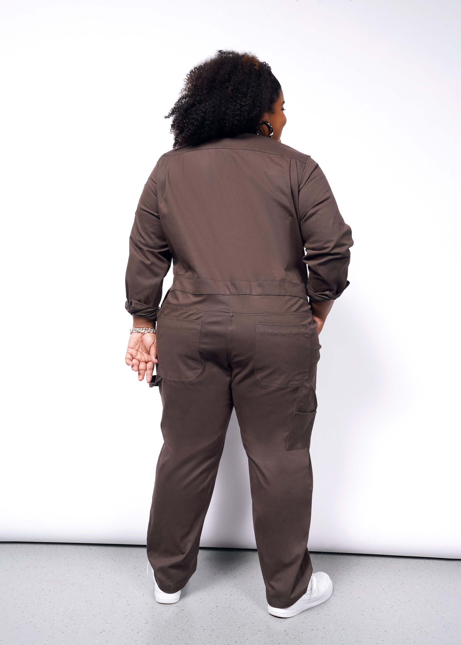 A person with curly hair faces away, wearing the oversized fit of The Essential Long Sleeve Coverall in brushed stretch cotton brown, paired with white sneakers. They stand against a white background.