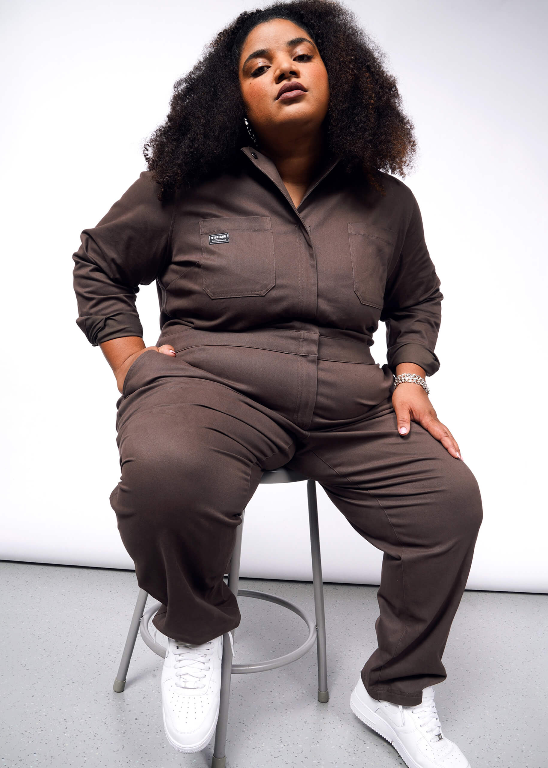 A person with curly hair and a confident expression sits on a stool, wearing The Essential Long Sleeve Coverall in an oversized fit made from brushed stretch cotton, paired with white sneakers. The background is plain white.