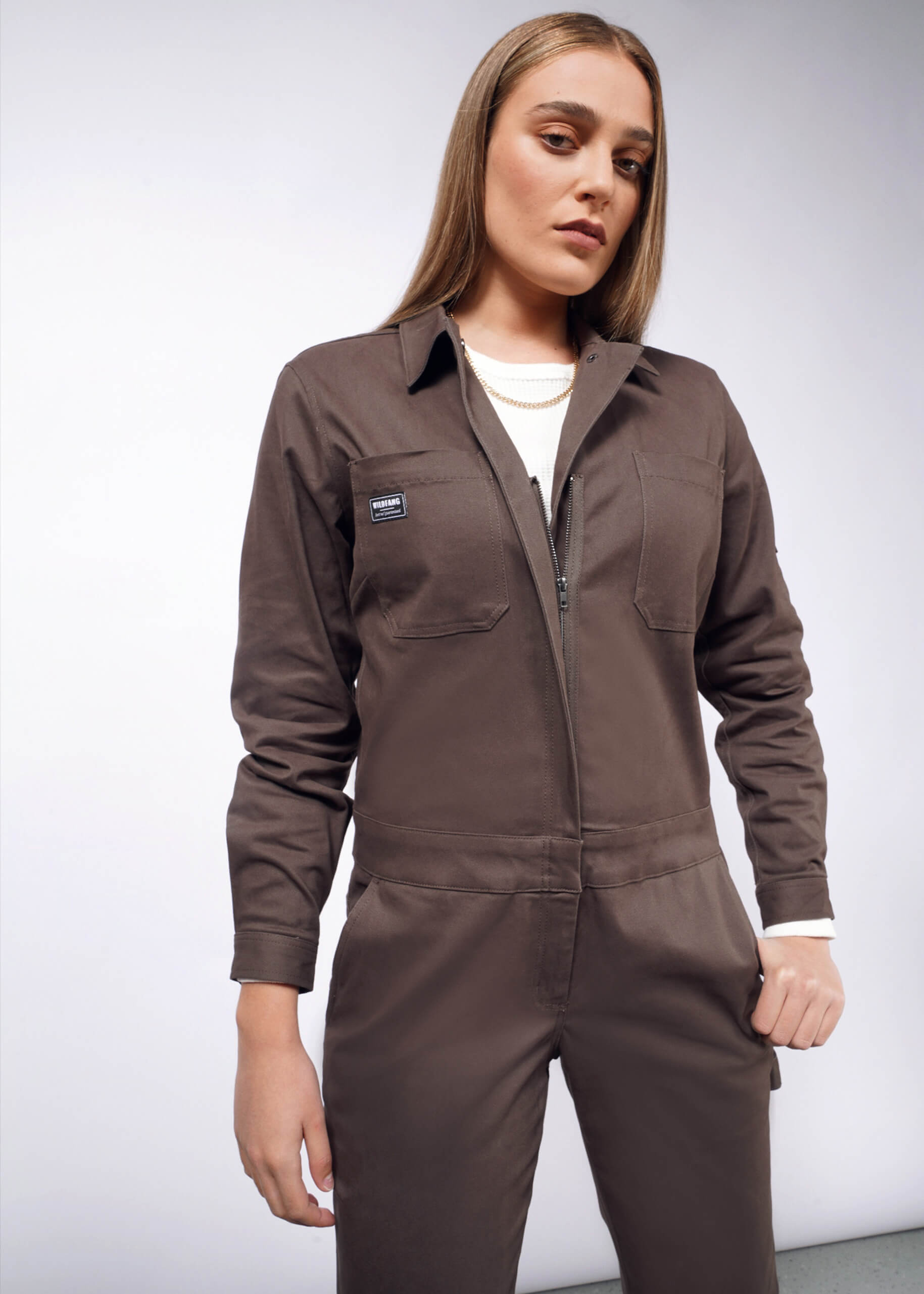 A woman with long hair wears The Essential Long Sleeve Coverall in dark brown brushed stretch cotton, paired with a white shirt underneath. It features two front pockets and a stylish zipper as she stands confidently against a plain background.