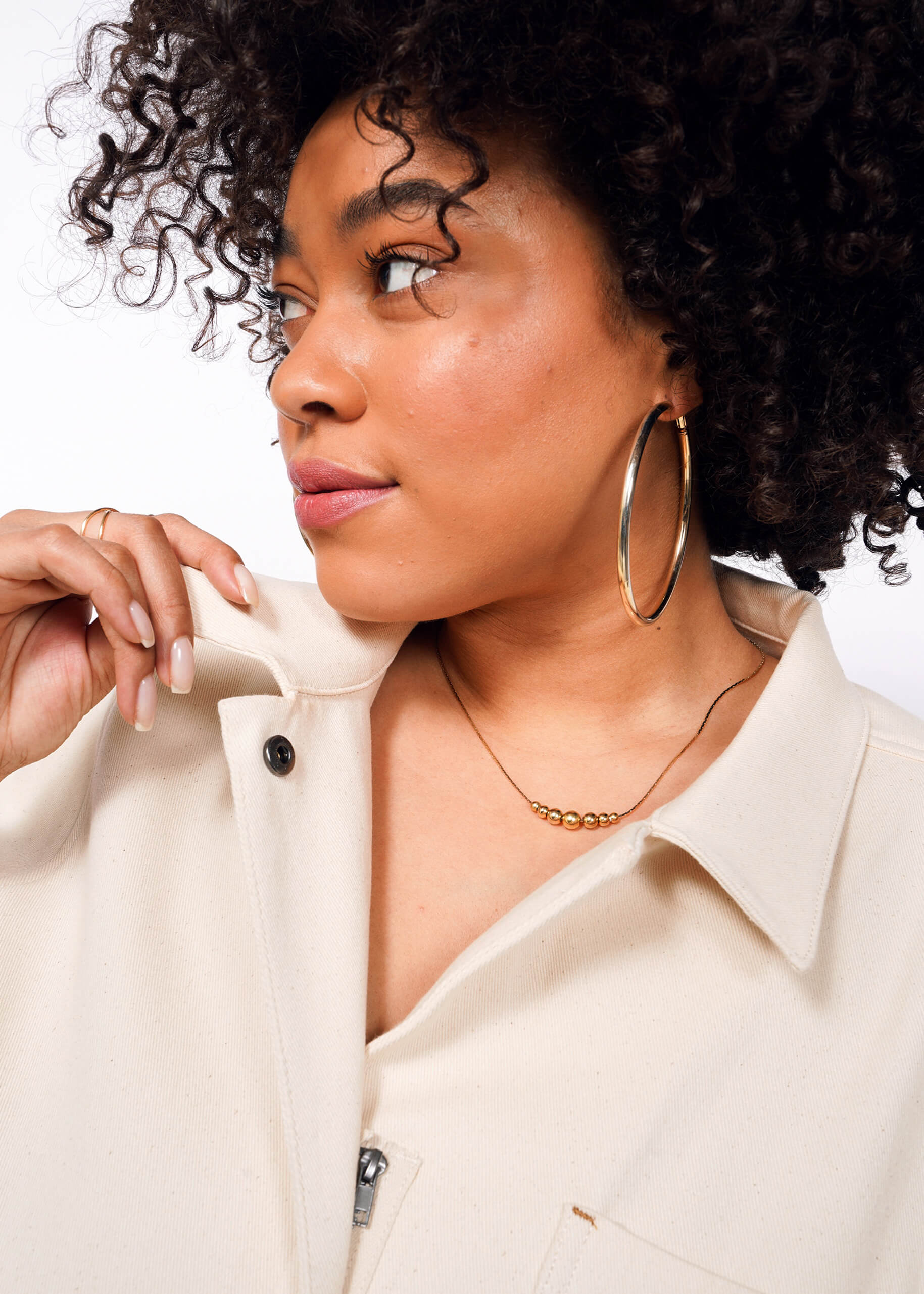 A person with curly hair and large hoop earrings looks to the side in a cream-colored jacket. One hand holds the collar, revealing a delicate necklace. The outfit is complemented by The Essential Denim High Waisted Coverall, set against a plain white backdrop.