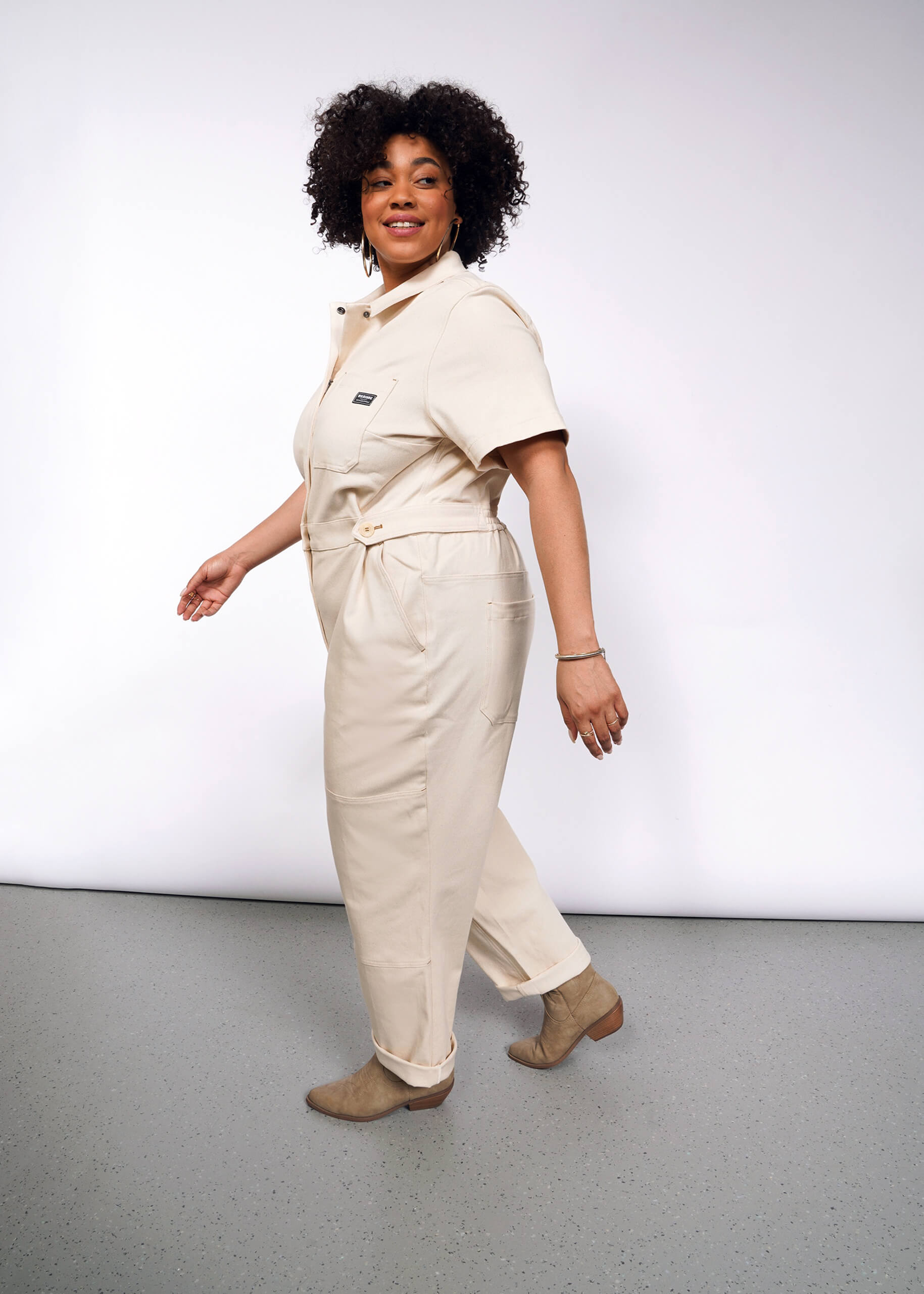 Someone with curly hair is dressed in The Essential Denim High Waisted Coverall featuring deep pockets and tan ankle boots. They stand on a gray floor, slightly turned with one foot forward, smiling against a plain white background.