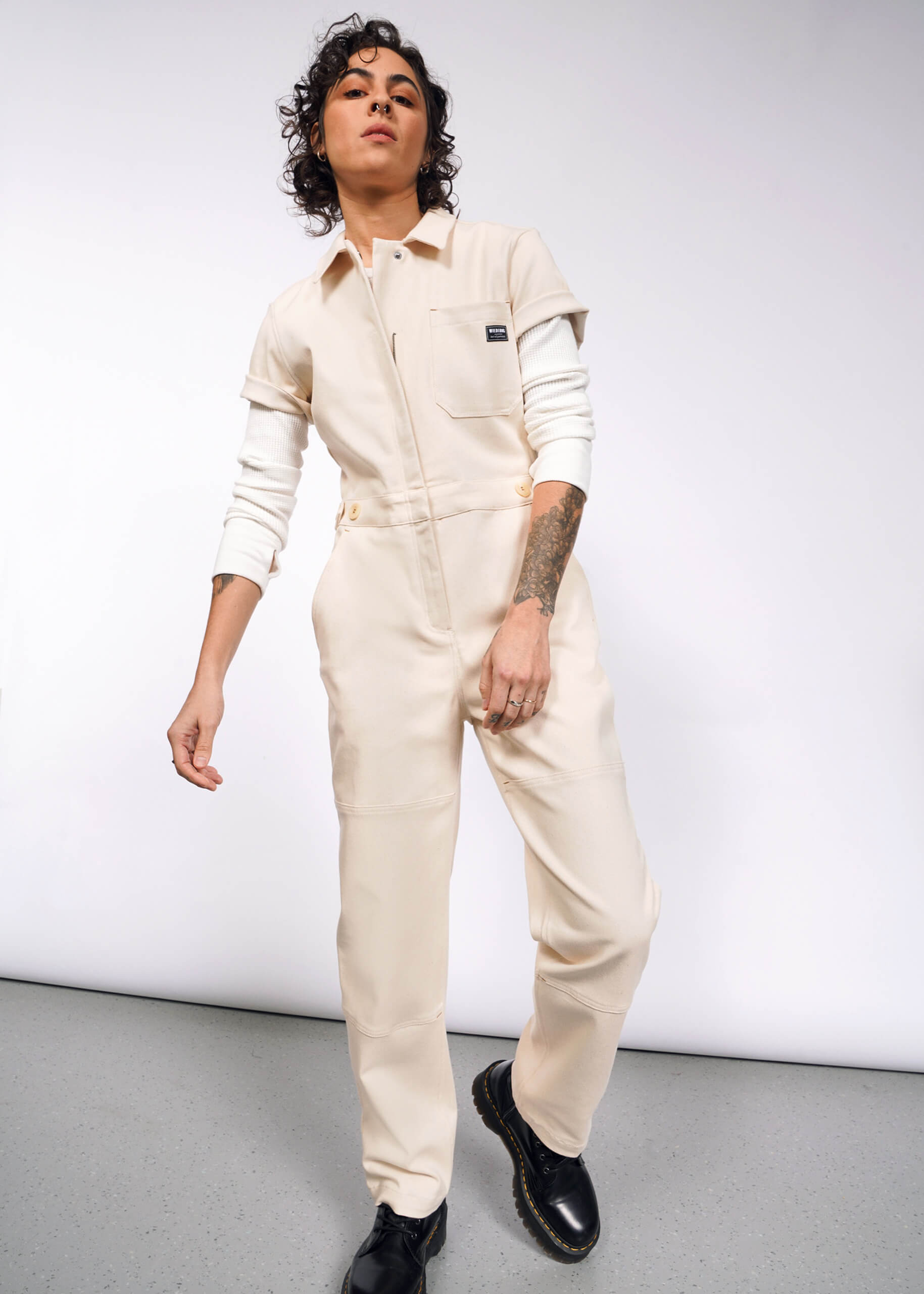 A person with curly hair confidently poses in The Essential Denim High Waisted Coverall over a white long-sleeve shirt, showcasing tattoos on one arm, against a plain background.