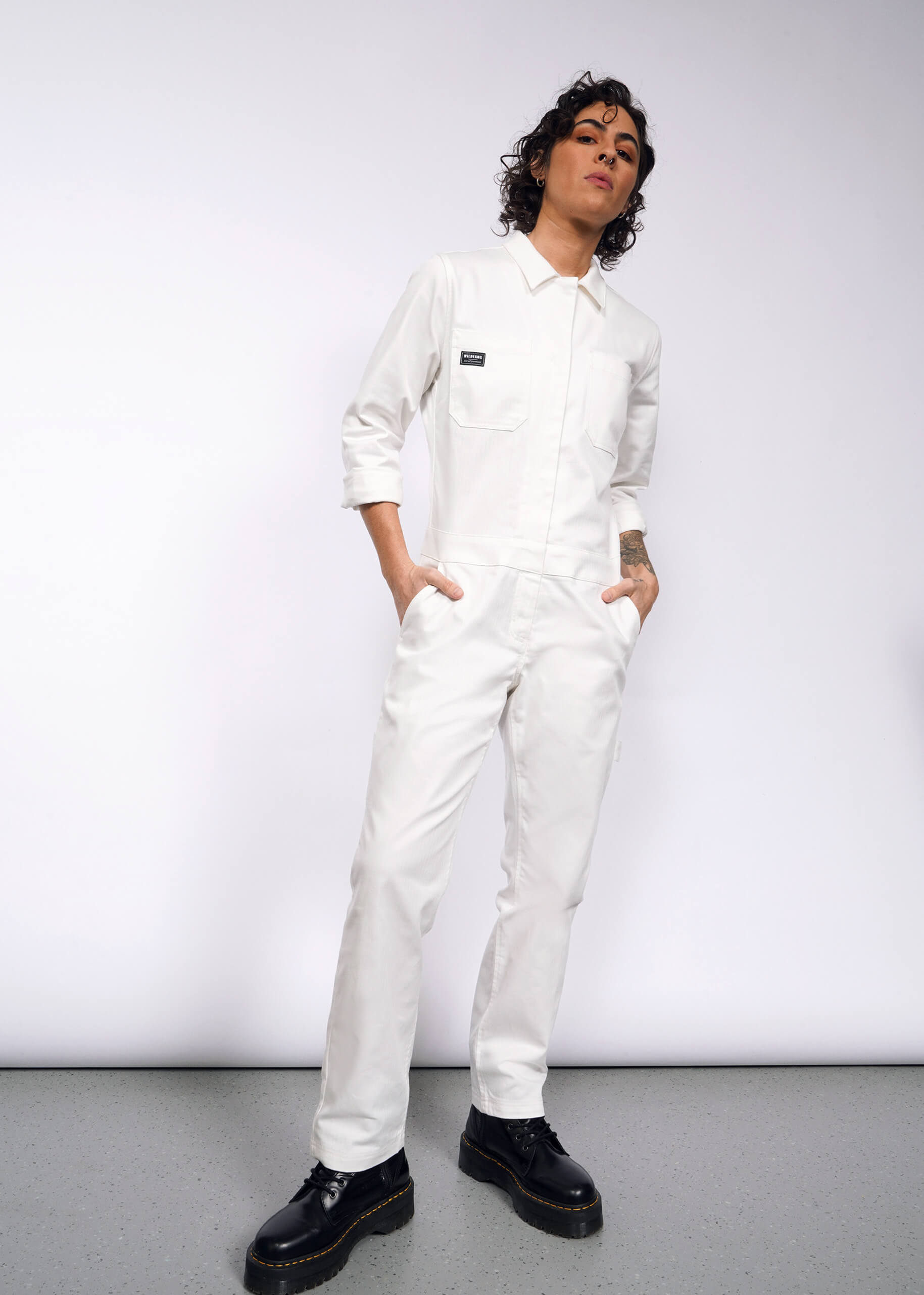 Sporting The Essential Long Sleeve Herringbone Coverall in white, a person with curly hair stands confidently against a plain backdrop. Featuring a full-length zipper and 10 pockets, the jumpsuit is paired with black shoes. Their hands rest in one pocket, and they gaze at the camera.