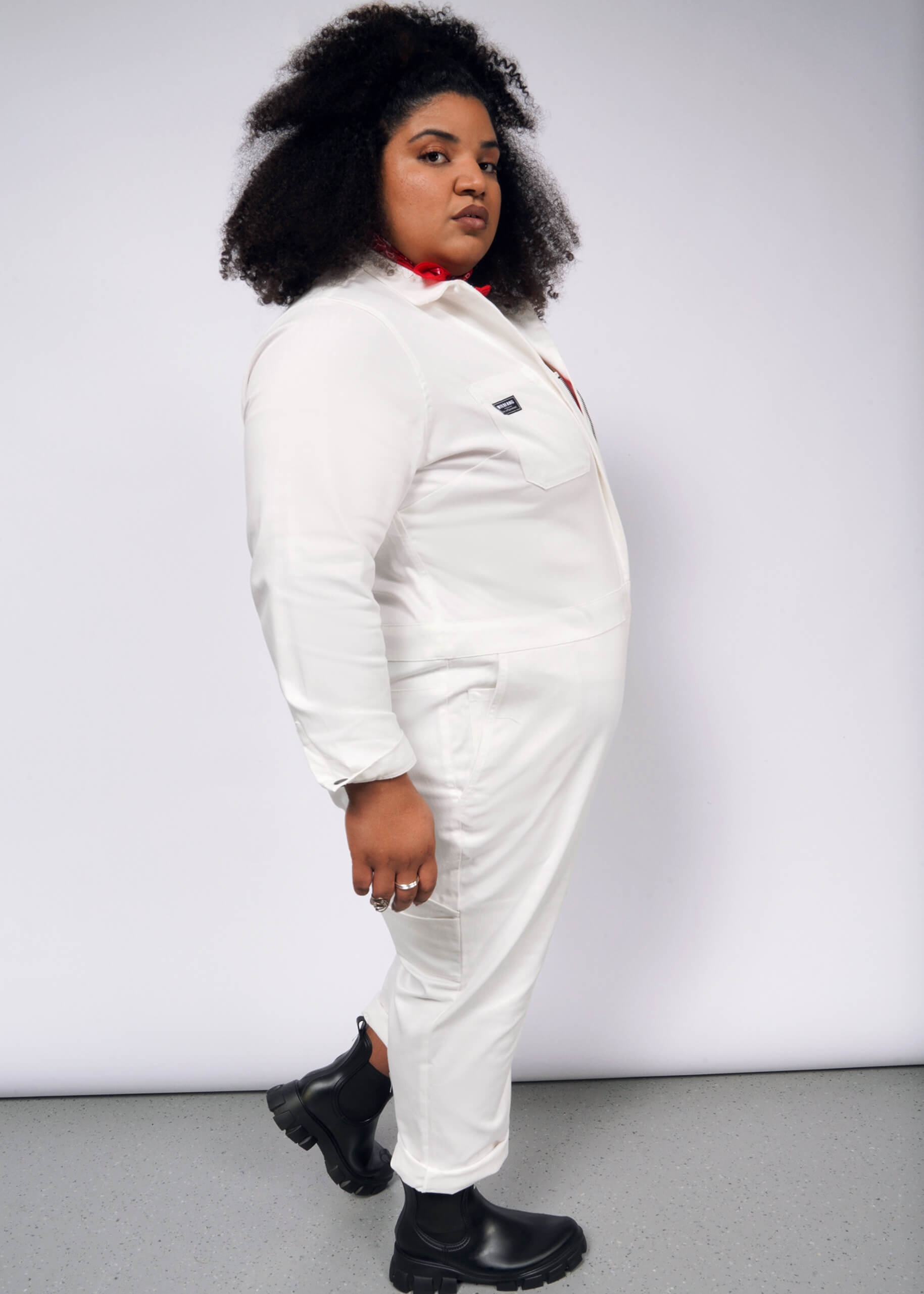 A person in The Essential Long Sleeve Herringbone Coverall, styled with black boots and a red scarf, stands against a gray background. Their curly hair frames their face as they look sideways, adding brightness to the minimalist look.