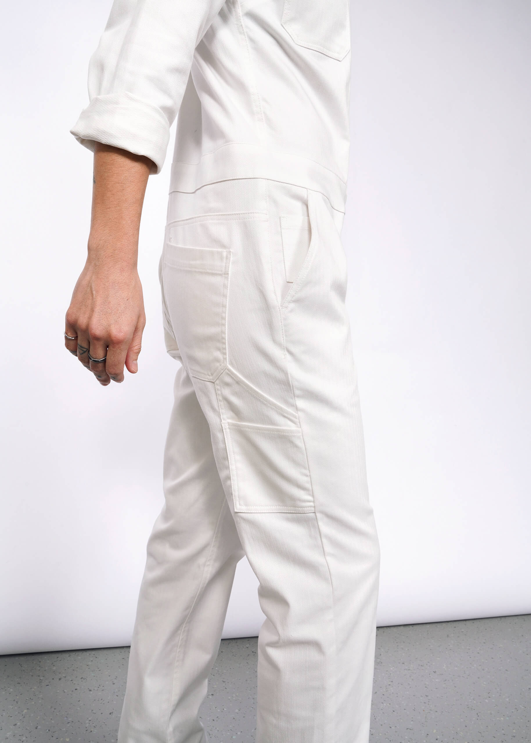 A person in The Essential Long Sleeve Herringbone Coverall, made from white herringbone cotton and featuring a full-length zipper and 10 pockets, stands against a light gray background. They are shown from the shoulder down with their arm slightly bent.