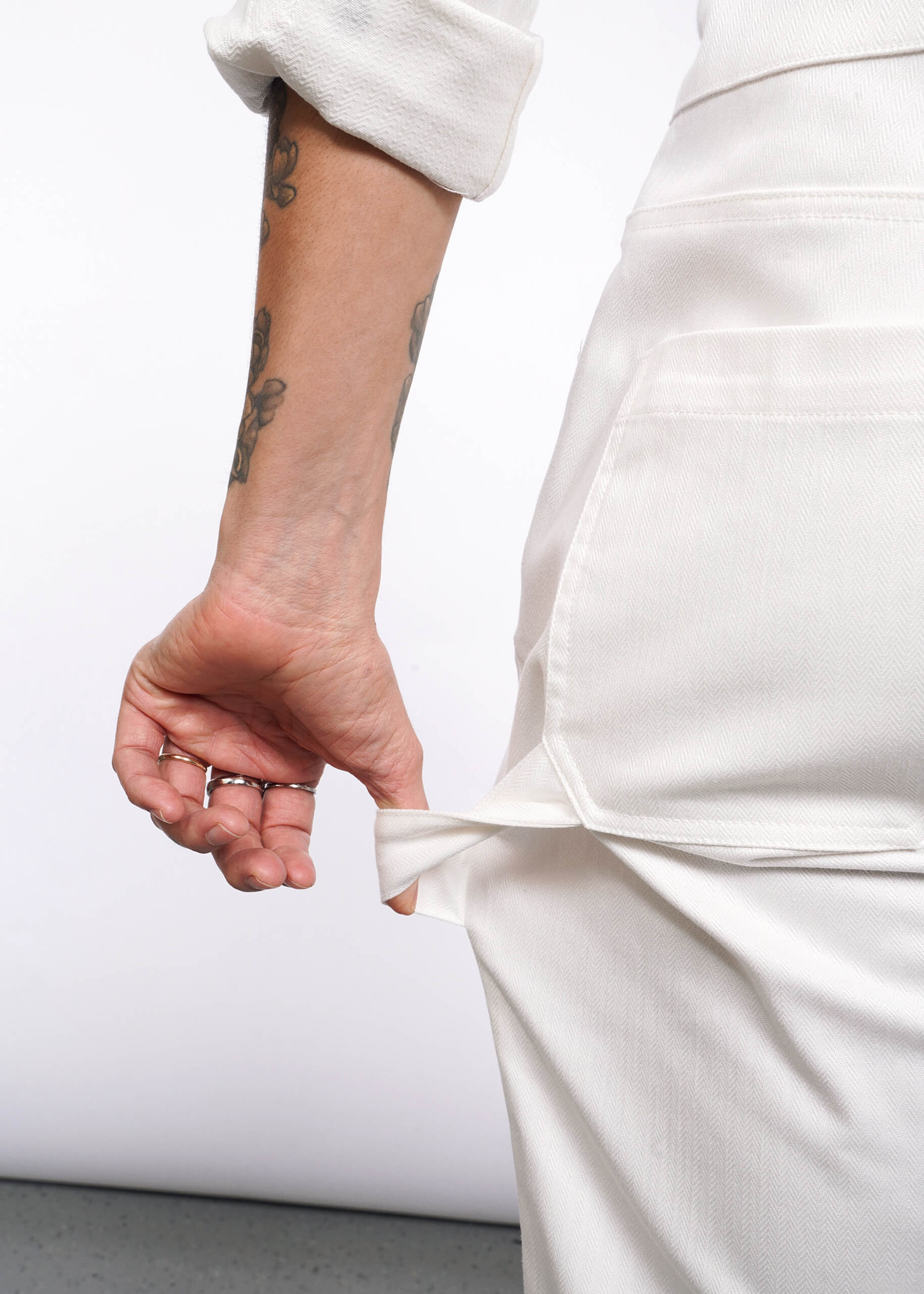 A tattooed person wearing The Essential Long Sleeve Herringbone Coverall in white, with the sleeve rolled up, shows the inside of an empty pocket. Their forearm and hand with rings appear against a plain white background.