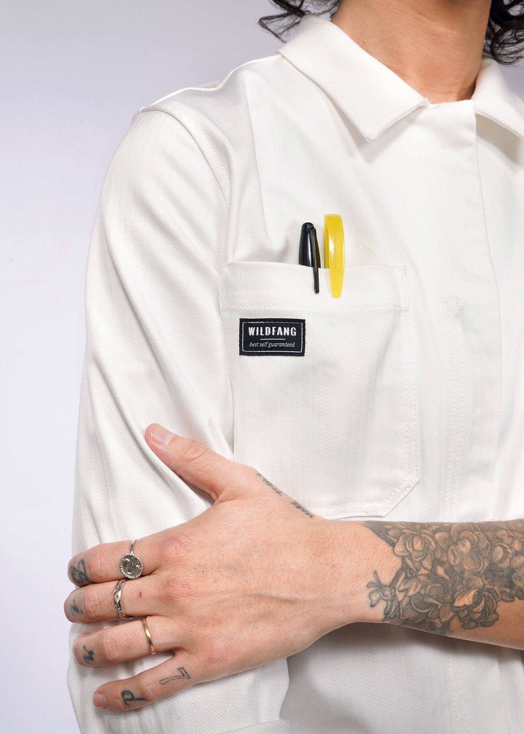 In a white Wildfang Essential Long Sleeve Herringbone Coverall, featuring herringbone cotton and 10 pockets, a person stands confidently. Pens peek from one pocket. Tattoos cover an arm, while gleaming rings adorn fingers as they cross arms in front of their body.