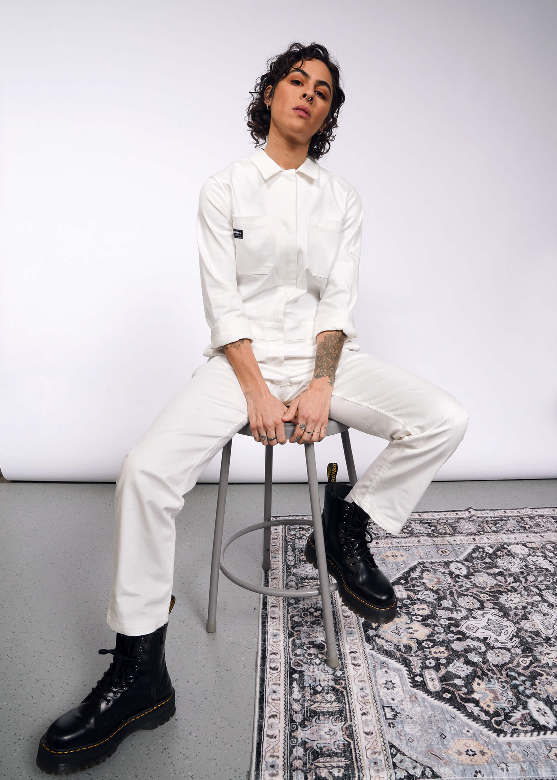 A curly-haired person sits on a stool in The Essential Long Sleeve Herringbone Coverall with a full-length zipper and black boots. Their relaxed arms rest on their knees against a plain backdrop, with part of a patterned rug visible on the floor.