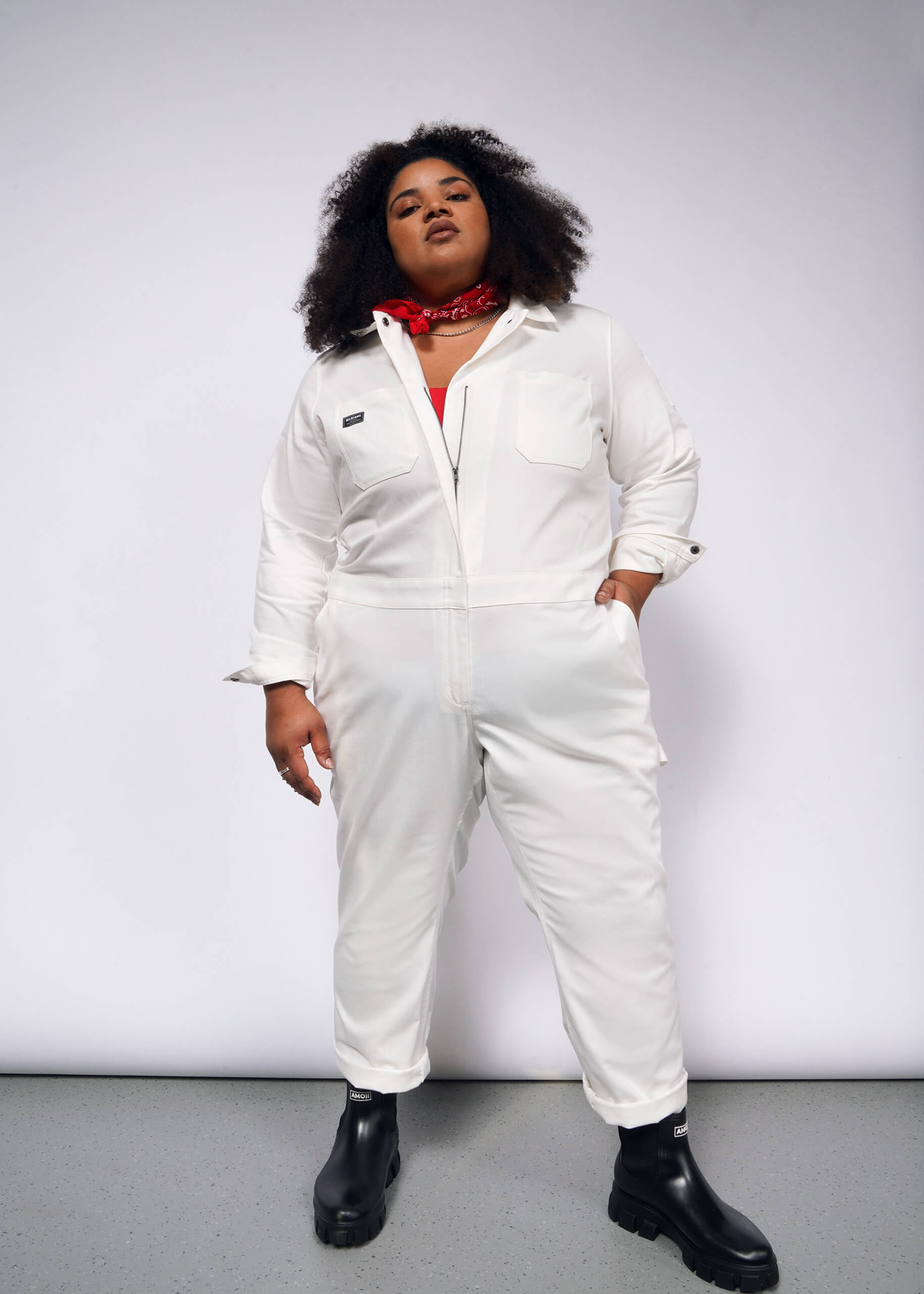 A curly-haired individual stands confidently against a plain background wearing The Essential Long Sleeve Herringbone Coverall, featuring a full-length zipper and multiple pockets for practicality, paired with black boots and accessorized with a red bandana around their neck.