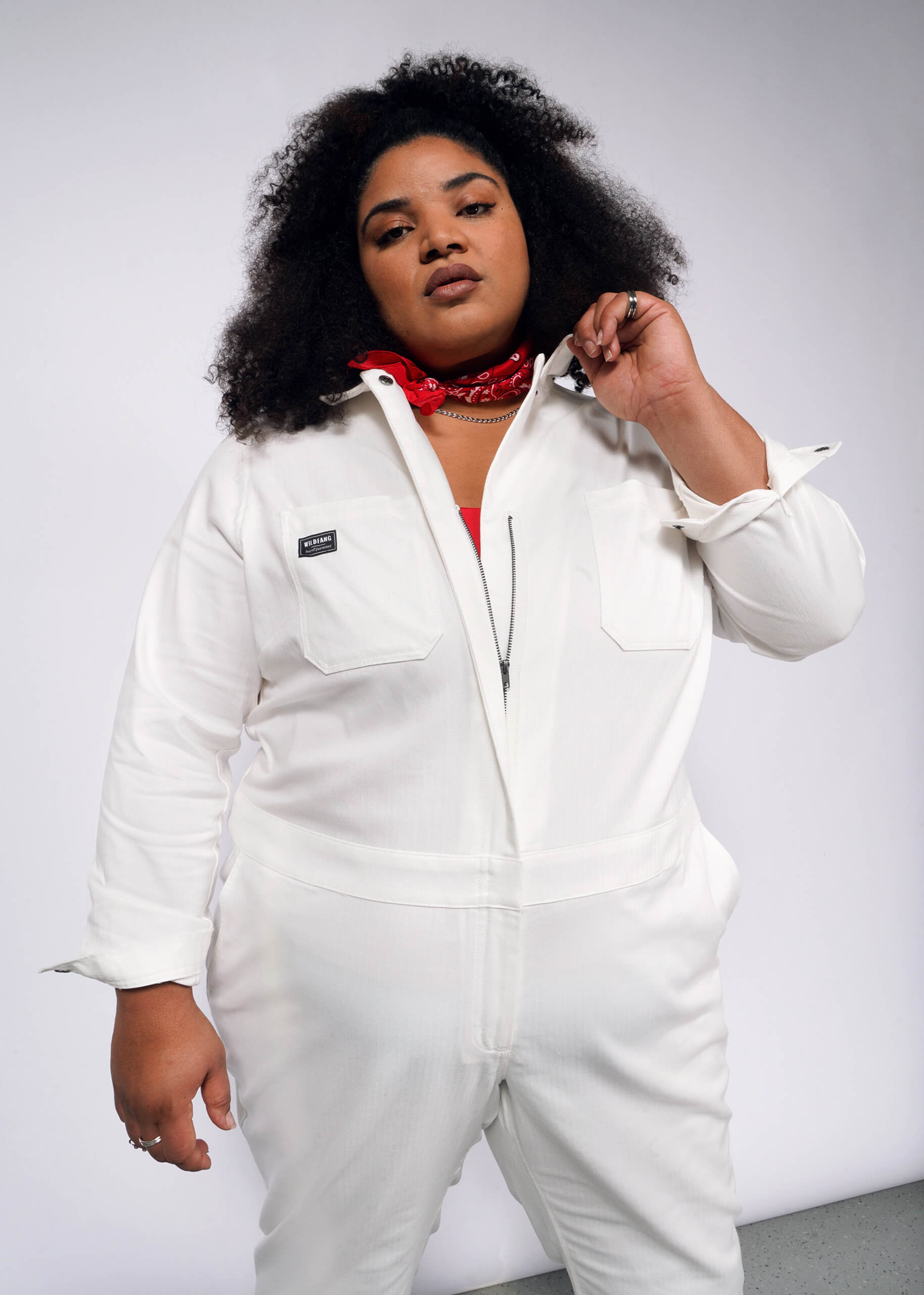 A person with curly hair wears The Essential Long Sleeve Herringbone Coverall, crafted from white cotton, and a red bandana around their neck. They adjust the full-length zipper of the coverall against a plain, light-colored background.