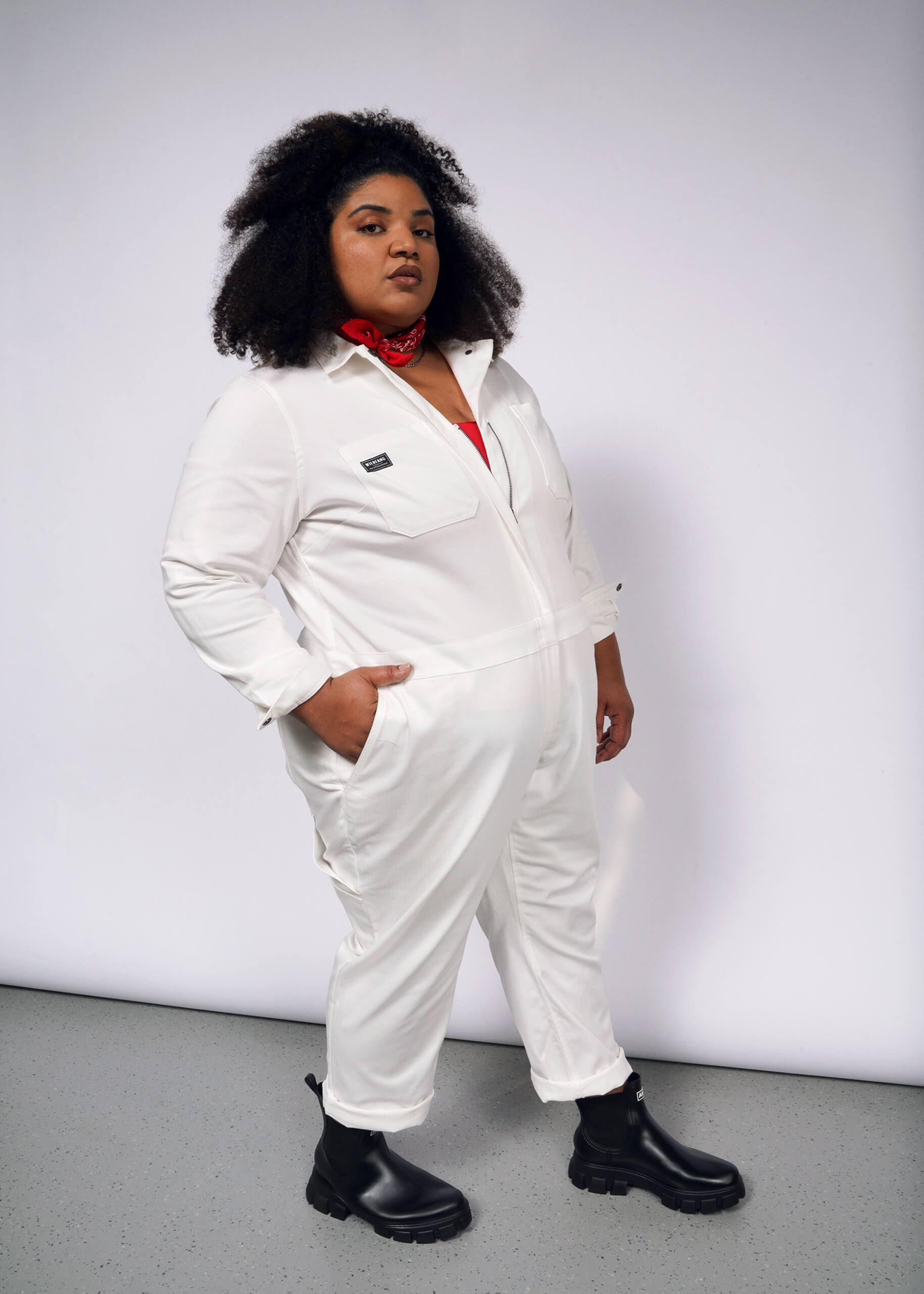 A person with curly hair stands confidently in The Essential Long Sleeve Herringbone Coverall, a white cotton jumpsuit with a full-length zipper. They wear black boots and a red bandana around their neck against a plain grey background.