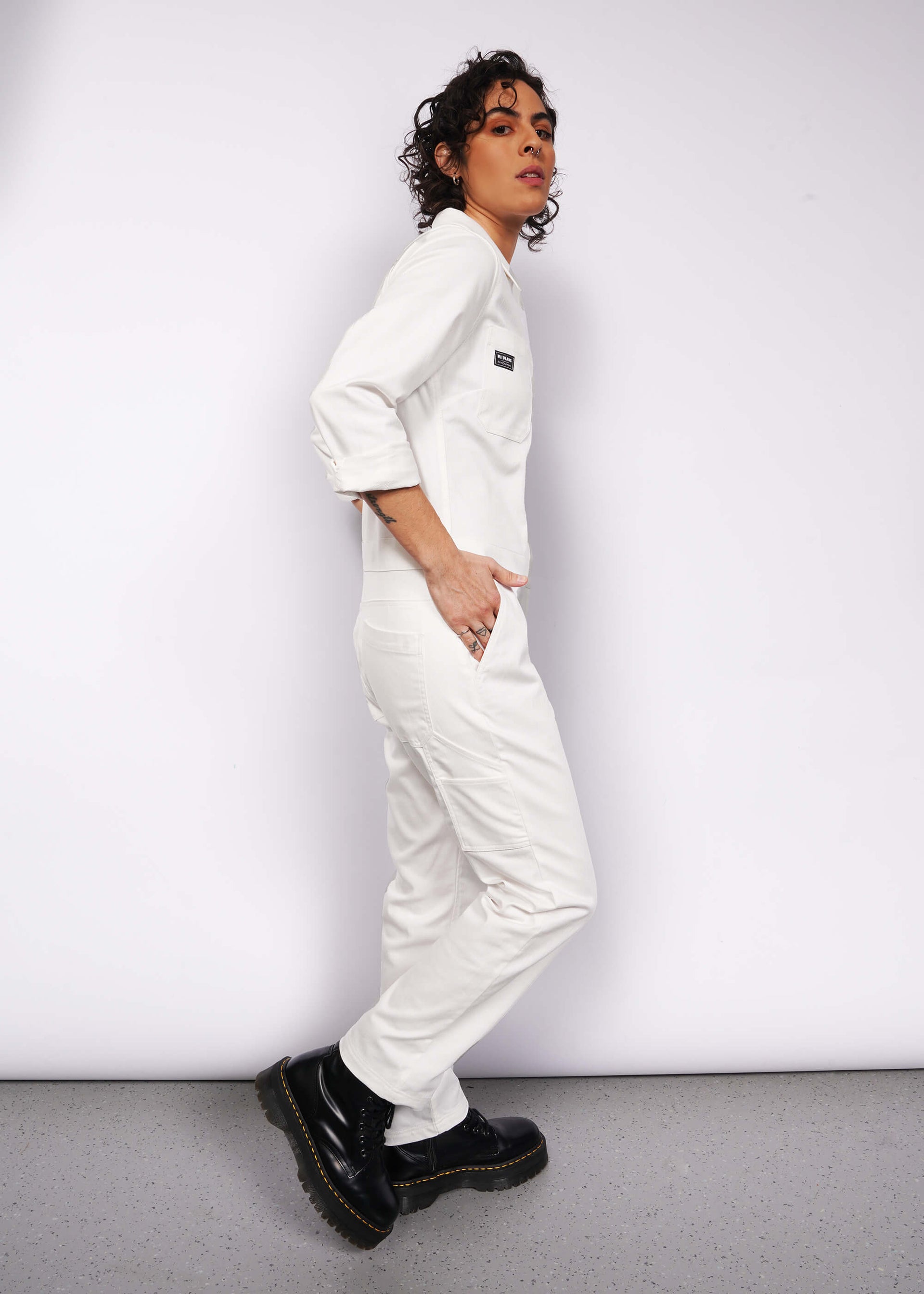 A person with short curly hair wears The Essential Long Sleeve Herringbone Coverall in white, paired with black boots, standing against a plain white wall. The coverall has a subtle logo, 10 pockets, and a full-length zipper. The gray speckled flooring lies beneath.