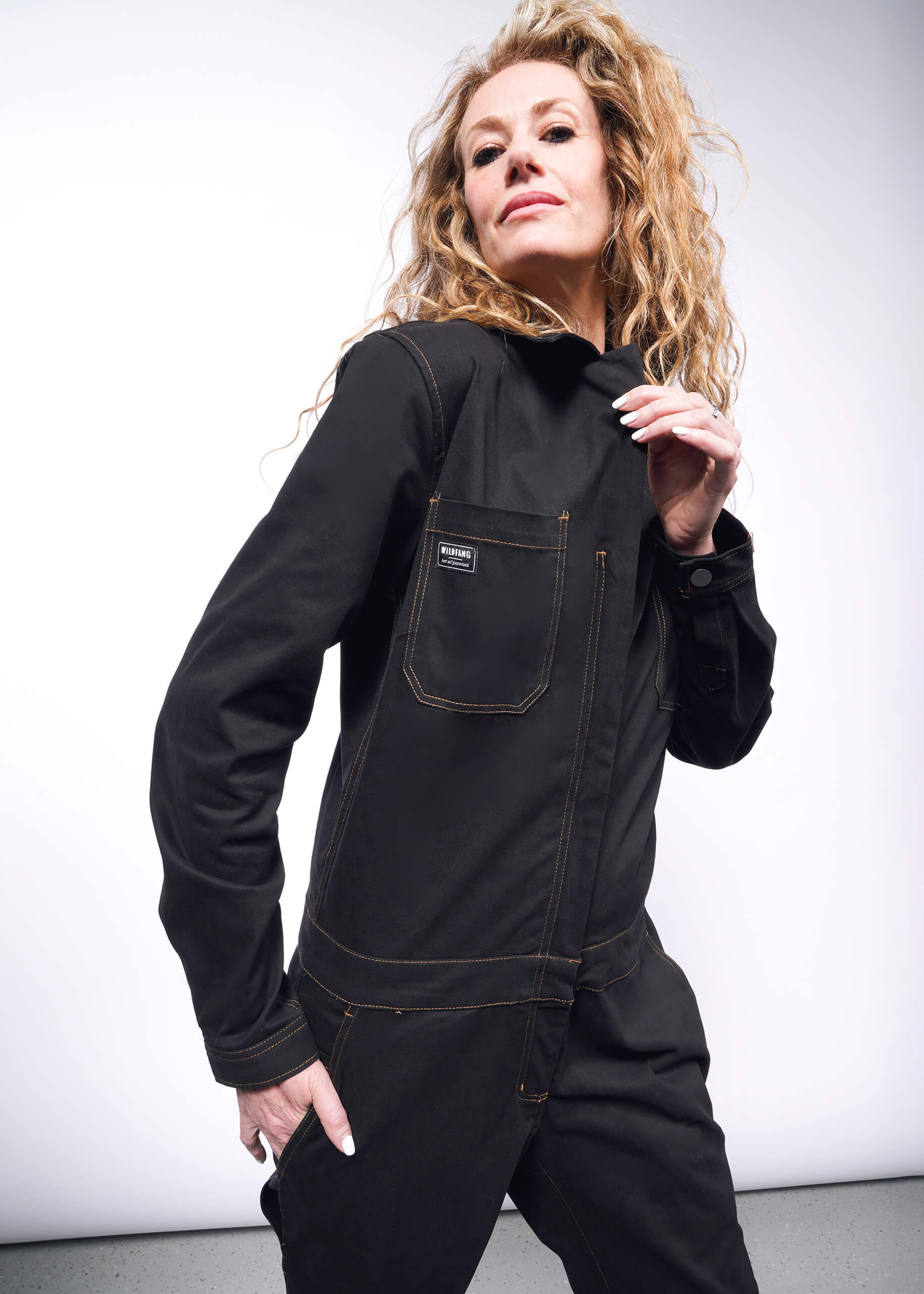 A person with long, curly hair confidently poses against a light background wearing The Essential Long Sleeve Coverall in black, made of brushed stretch cotton. This oversized outfit features visible stitching and numerous pockets.