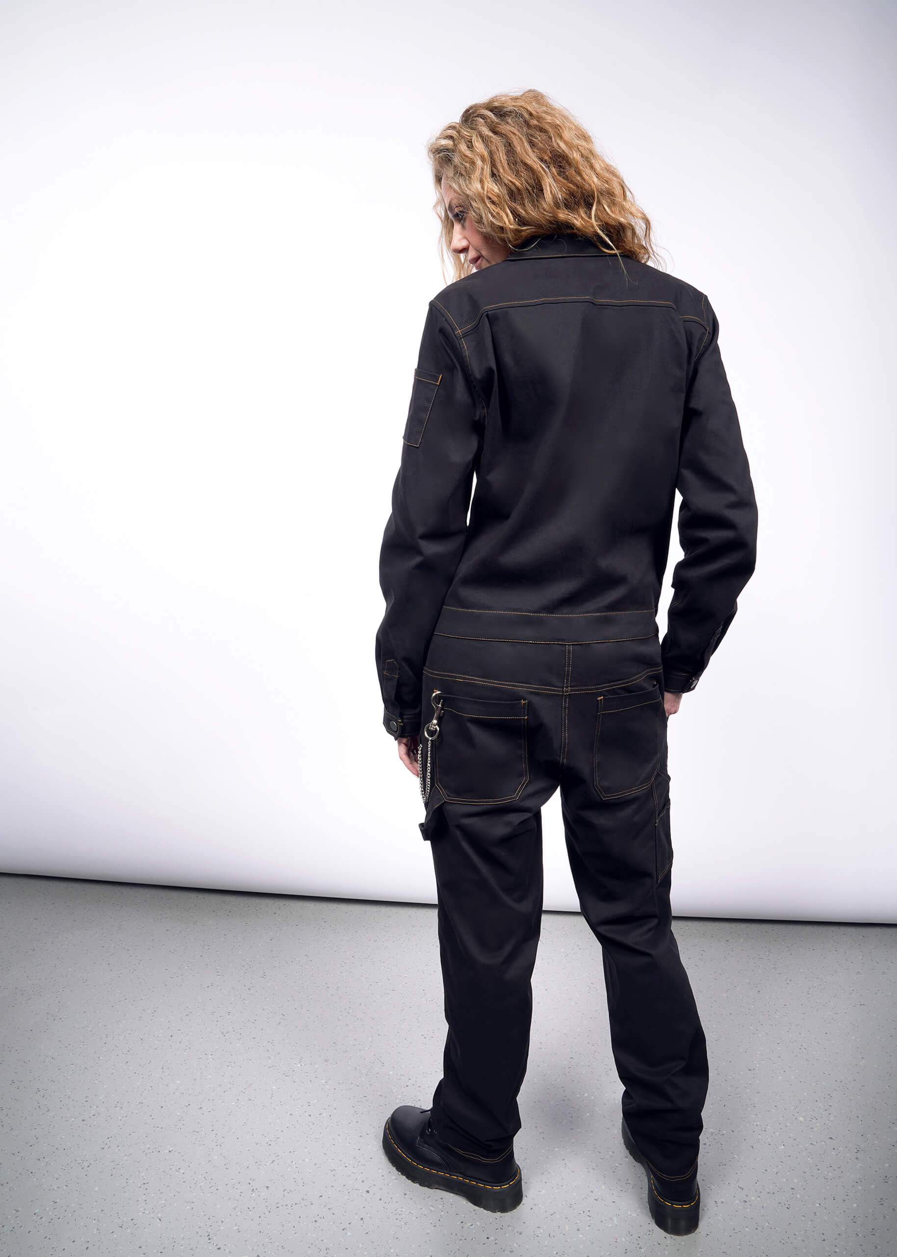 A person with long curly hair stands back to the camera, showcasing The Essential Long Sleeve Coverall in an oversized fit and rugged black boots. Crafted from brushed stretch cotton, it features 10 pockets and rests elegantly on the light gray floor of the well-lit room.