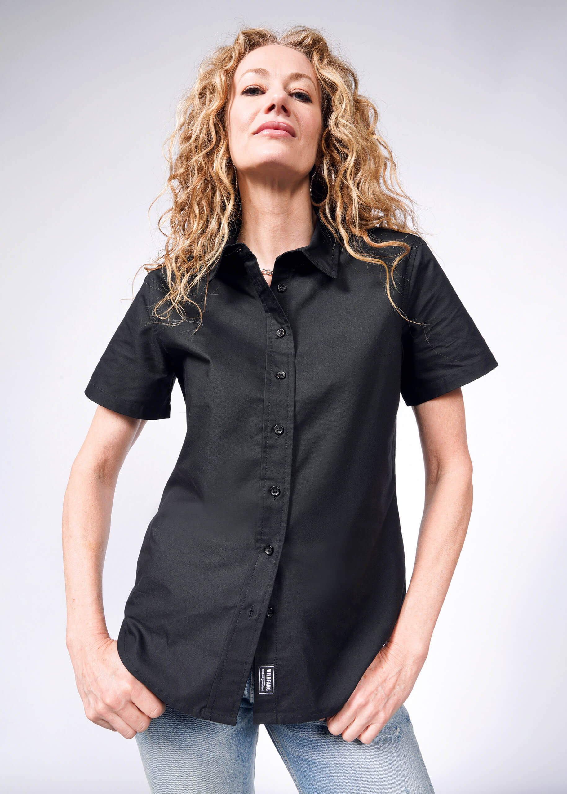 A person with long, curly hair stands confidently against a neutral background, wearing The Essential Oxford Button Up in black with branded buttons and light blue jeans.