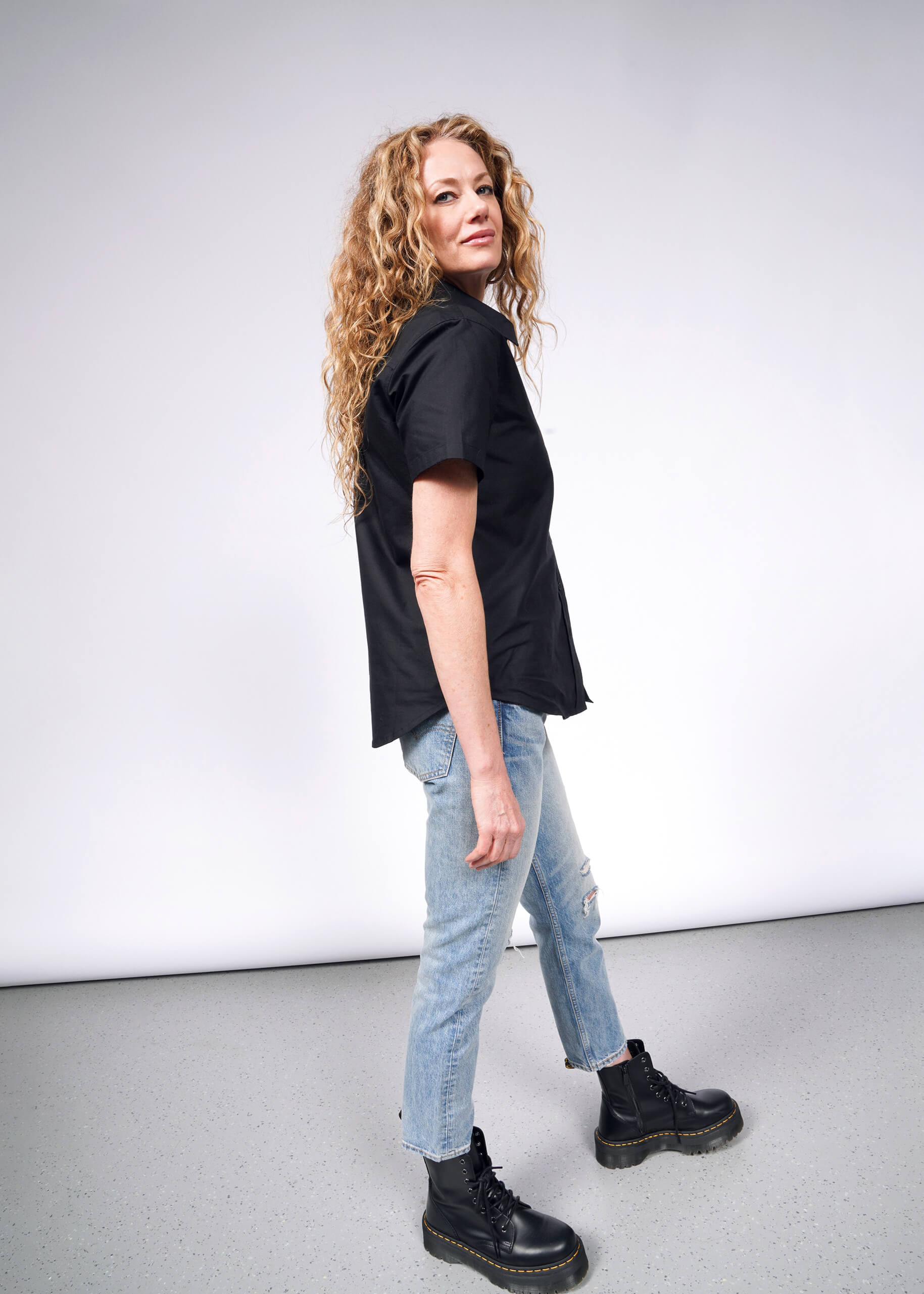 A woman with long curly hair stands in profile against a white background. She’s wearing a black top, ripped light blue jeans, and black boots, complemented by The Essential Oxford Button Up featuring Wildfang branded buttons for a tailored fit.