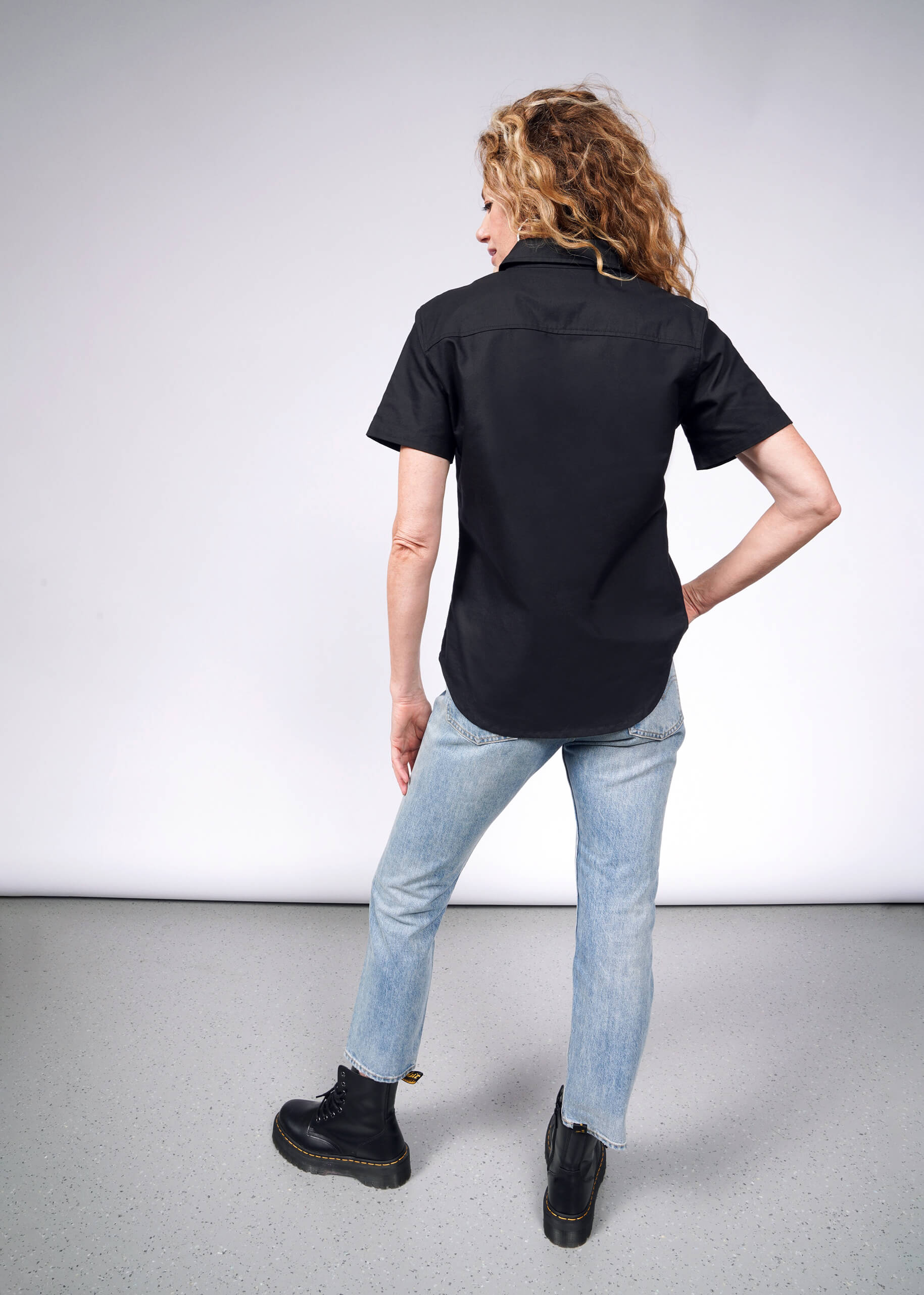 A woman with curly hair stands facing away, wearing The Essential Oxford Button Up in black, paired with light blue jeans and black boots. One hand rests on her hip in a neutral studio setting.