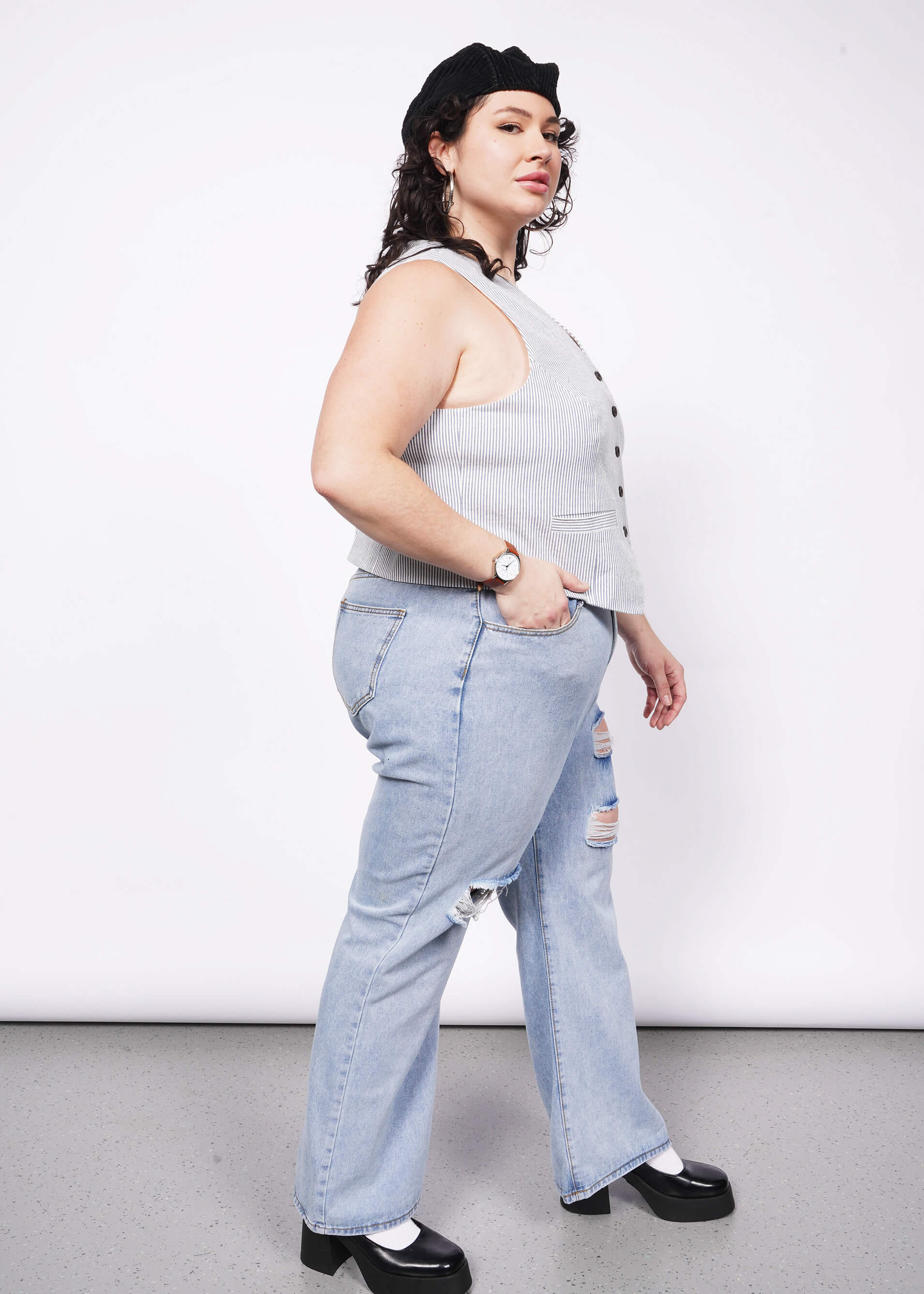 Person with short curly black hair looks at camera wearing seersucker blue and white vest and jeans.