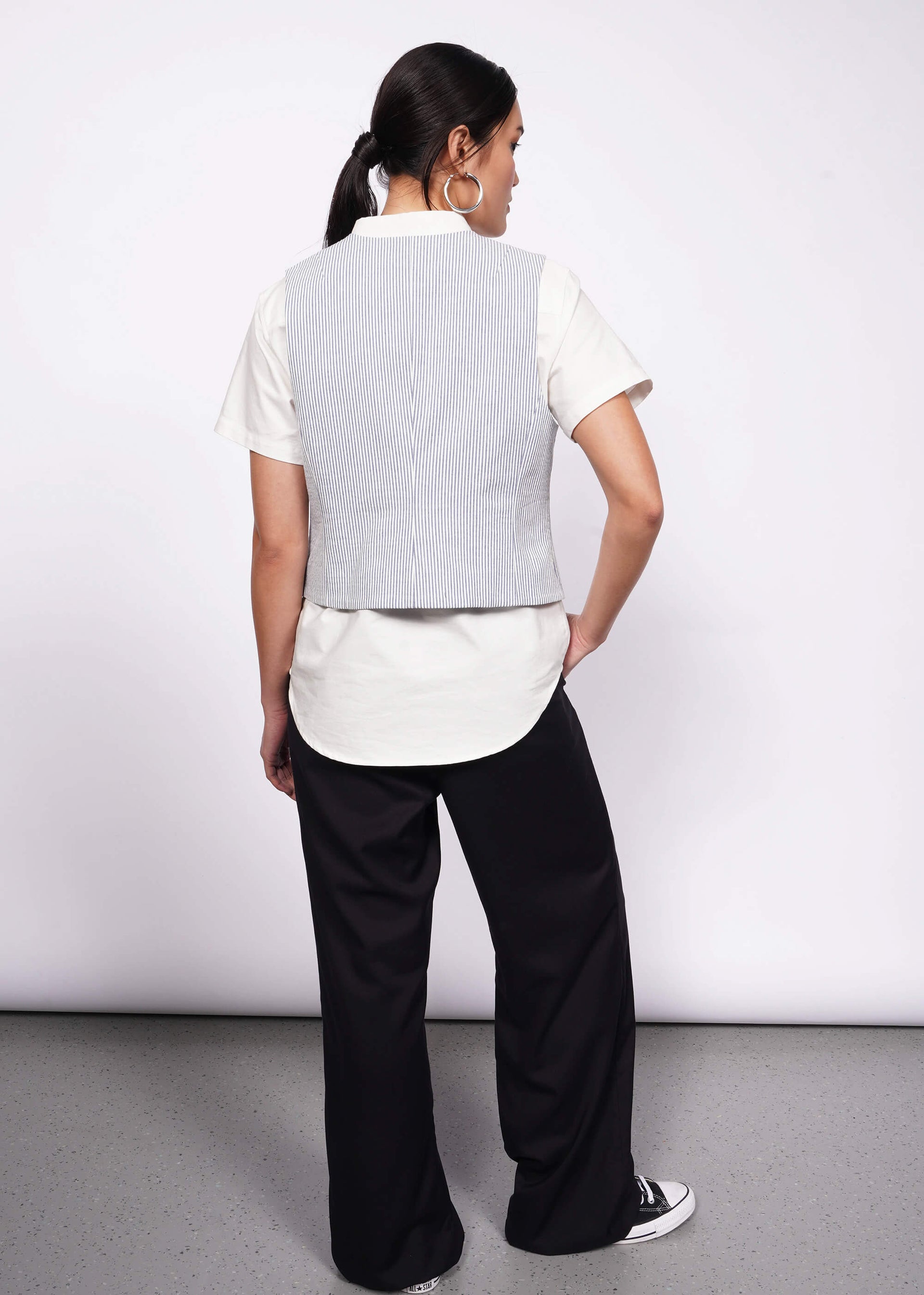 Back of person with tied back black hair wearing white short sleeve button up, a blue and white seersucker vest, black wide leg trouser, and Converse.