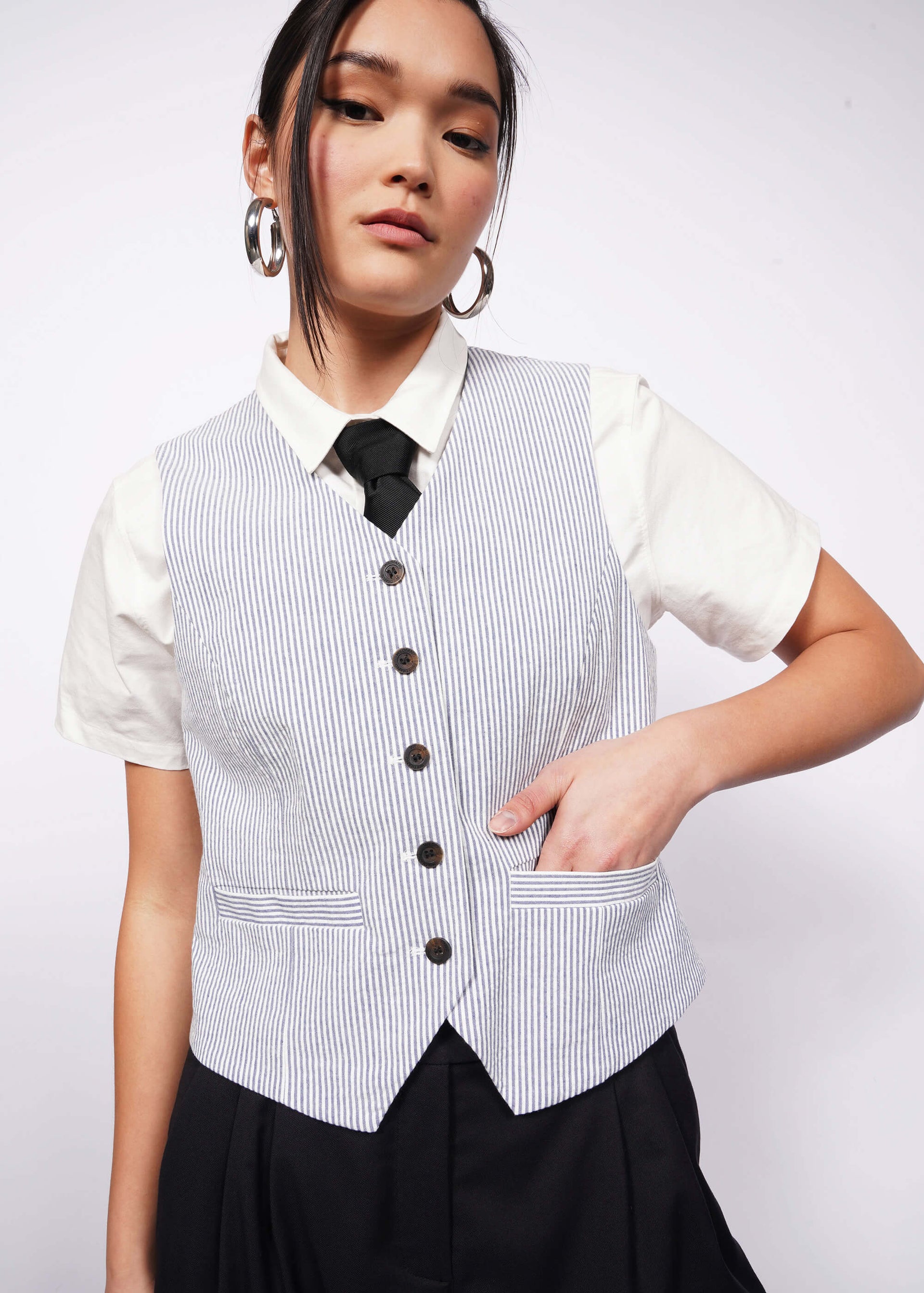 Person with tied back black hair wears white short sleeve button up, black tie, and blue and white seersucker vest.