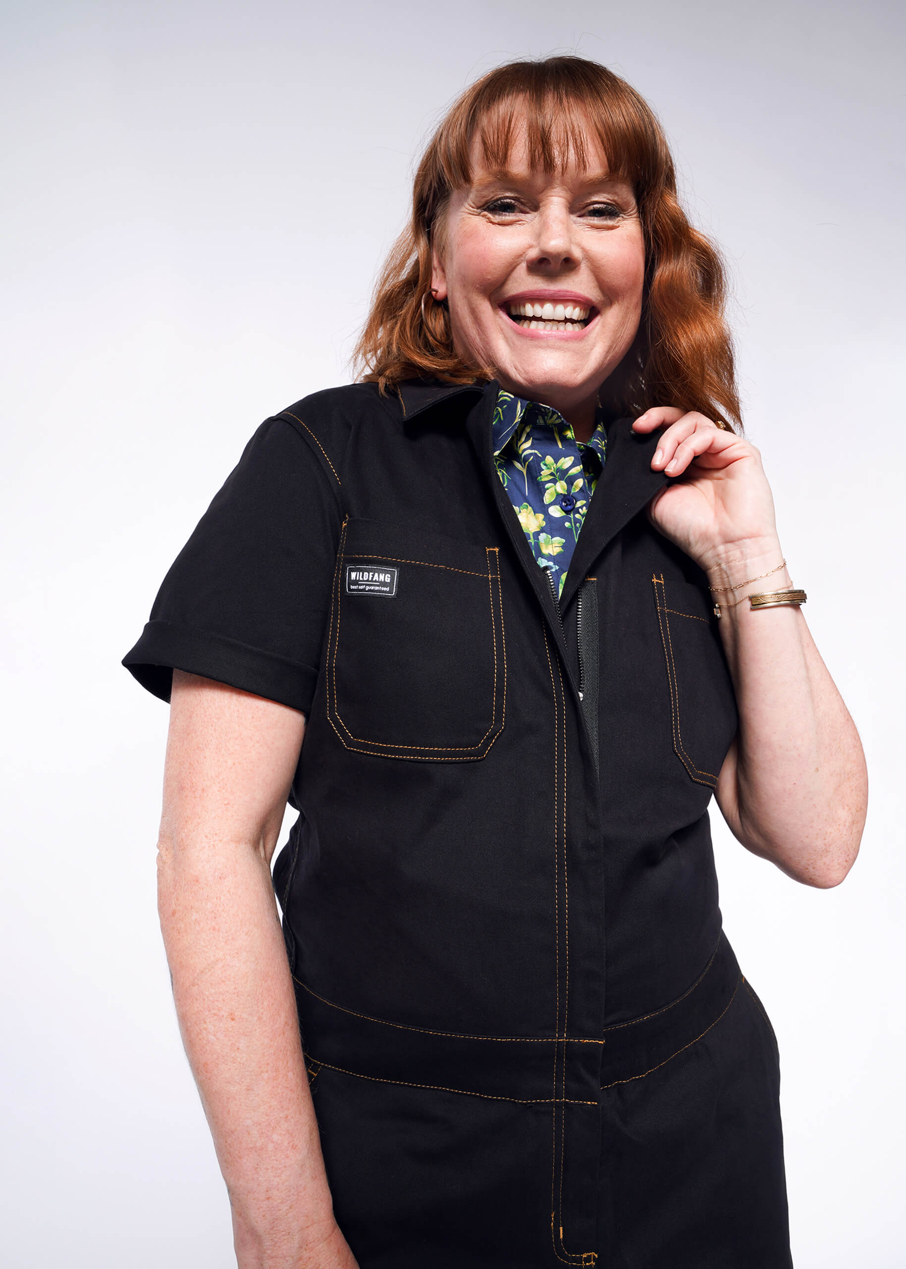 A person with shoulder-length brown hair smiles while wearing The Essential Coverall, a black oversized jumpsuit with yellow stitching and a floral shirt underneath. Made of stretch cotton, it features 9 pockets and theyre posed against a plain white background.