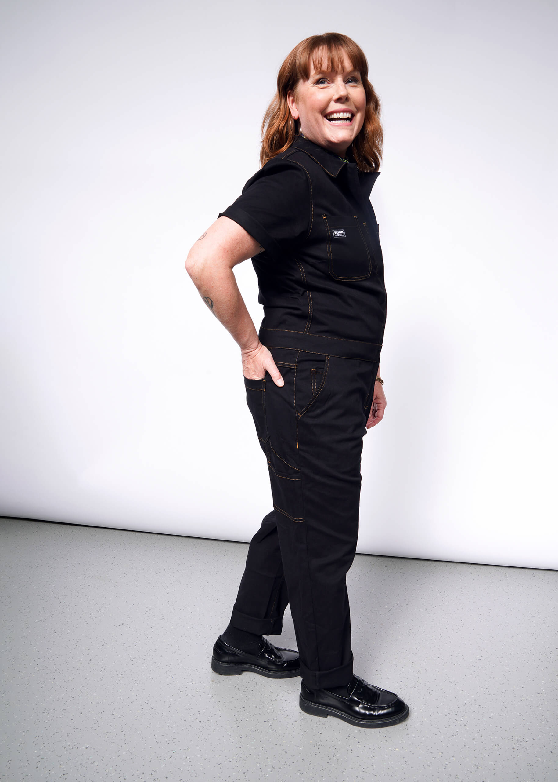 A person stands confidently against a plain background, wearing The Essential Coverall—a black stretch cotton jumpsuit with 9 pockets—and shiny black shoes. With brown hair and a cheerful smile, they rest their hands on their hips.