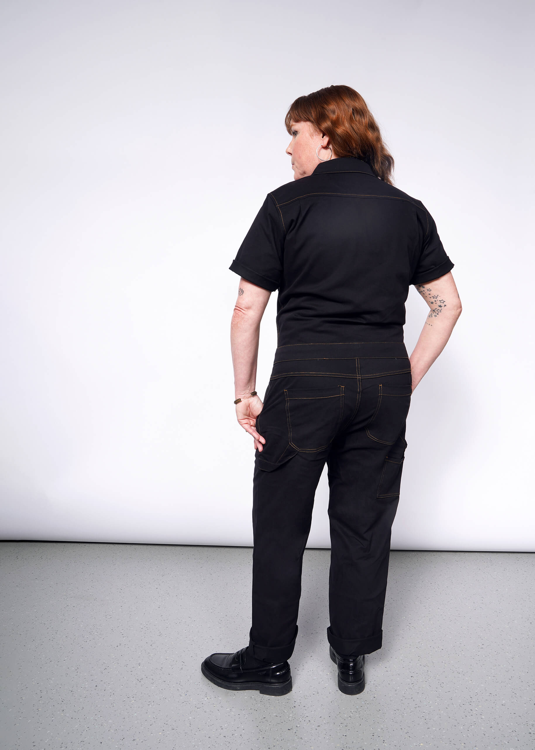 With medium-length brown hair, an individual is seen from behind wearing The Essential Coverall in black, standing in a light-colored room featuring a textured gray floor.