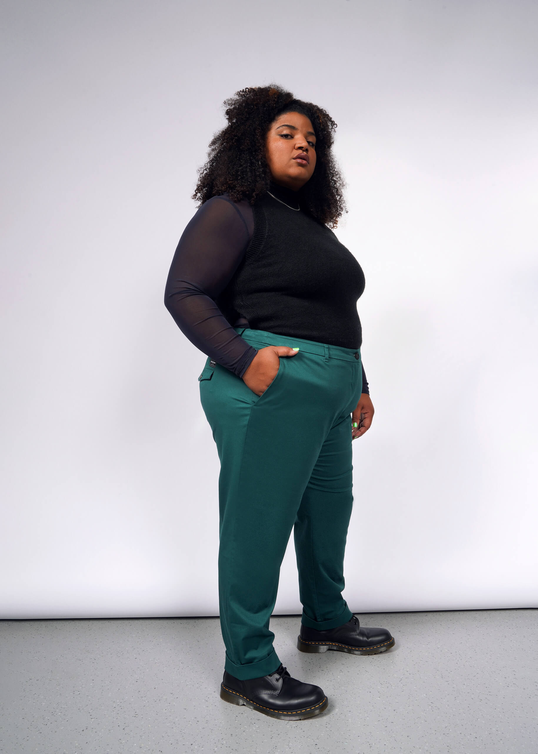 A person stands confidently against a white background, wearing a black top with sheer sleeves, The Essential Trouser in teal, and black boots. They have curly hair and their hands in their pockets.