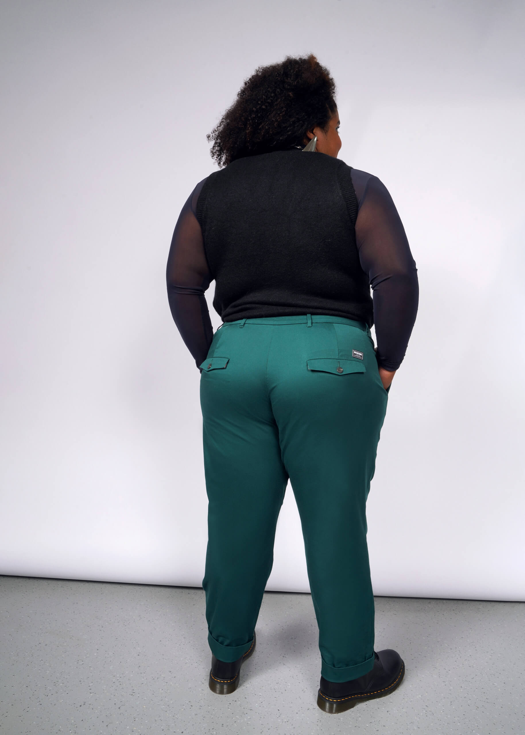 A person stands facing away, wearing a black sleeveless top with sheer sleeves and teal The Essential Trouser. They have short curly hair and hands in pockets, set against a white background, showcasing a wardrobe staple perfect for effortless style.