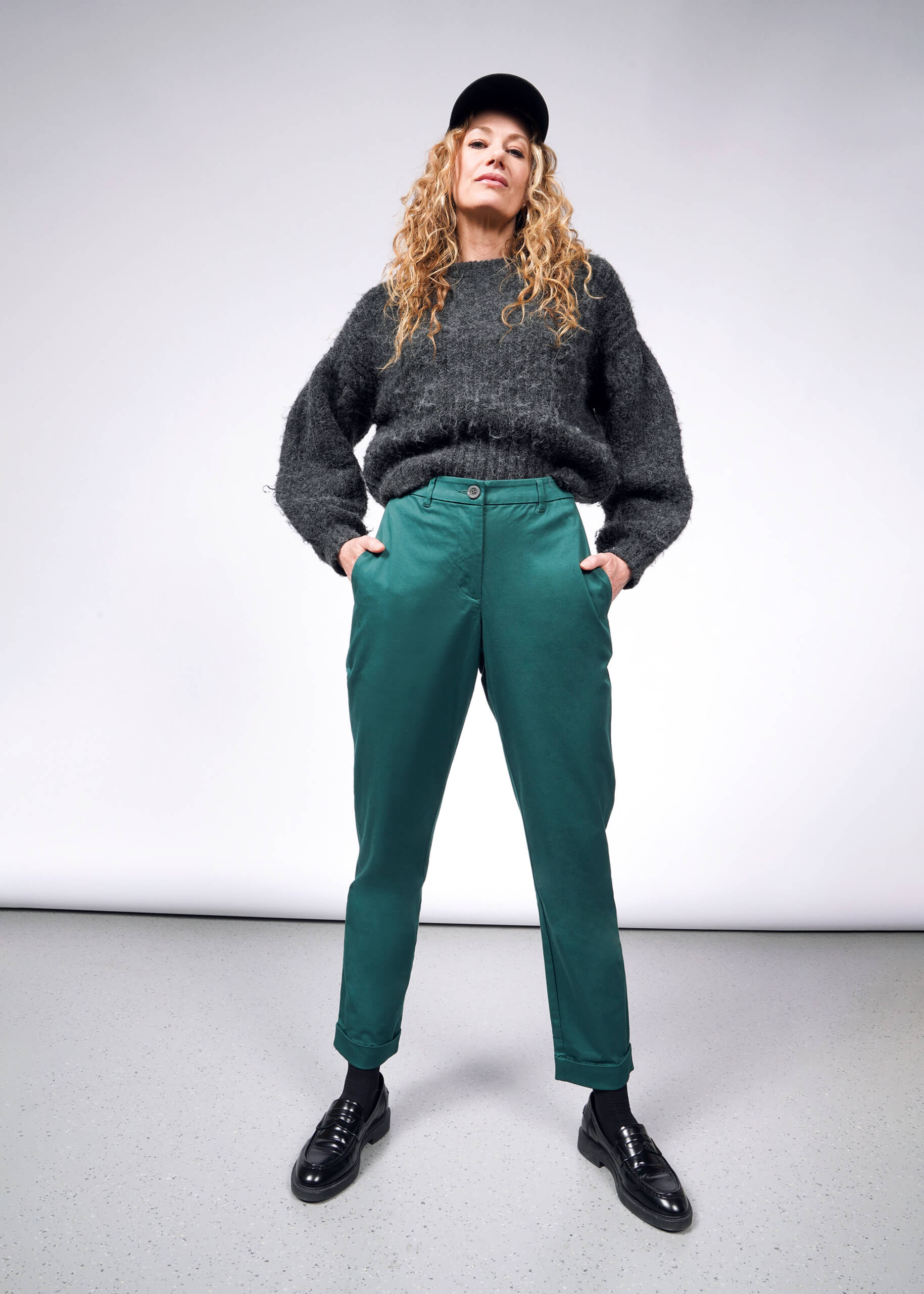 A person with long curly hair is wearing a black cap, gray sweater, and The Essential Trouser in green, paired with black shoes. They stand confidently with hands in pockets against a plain background, showcasing their flair for wardrobe staples.