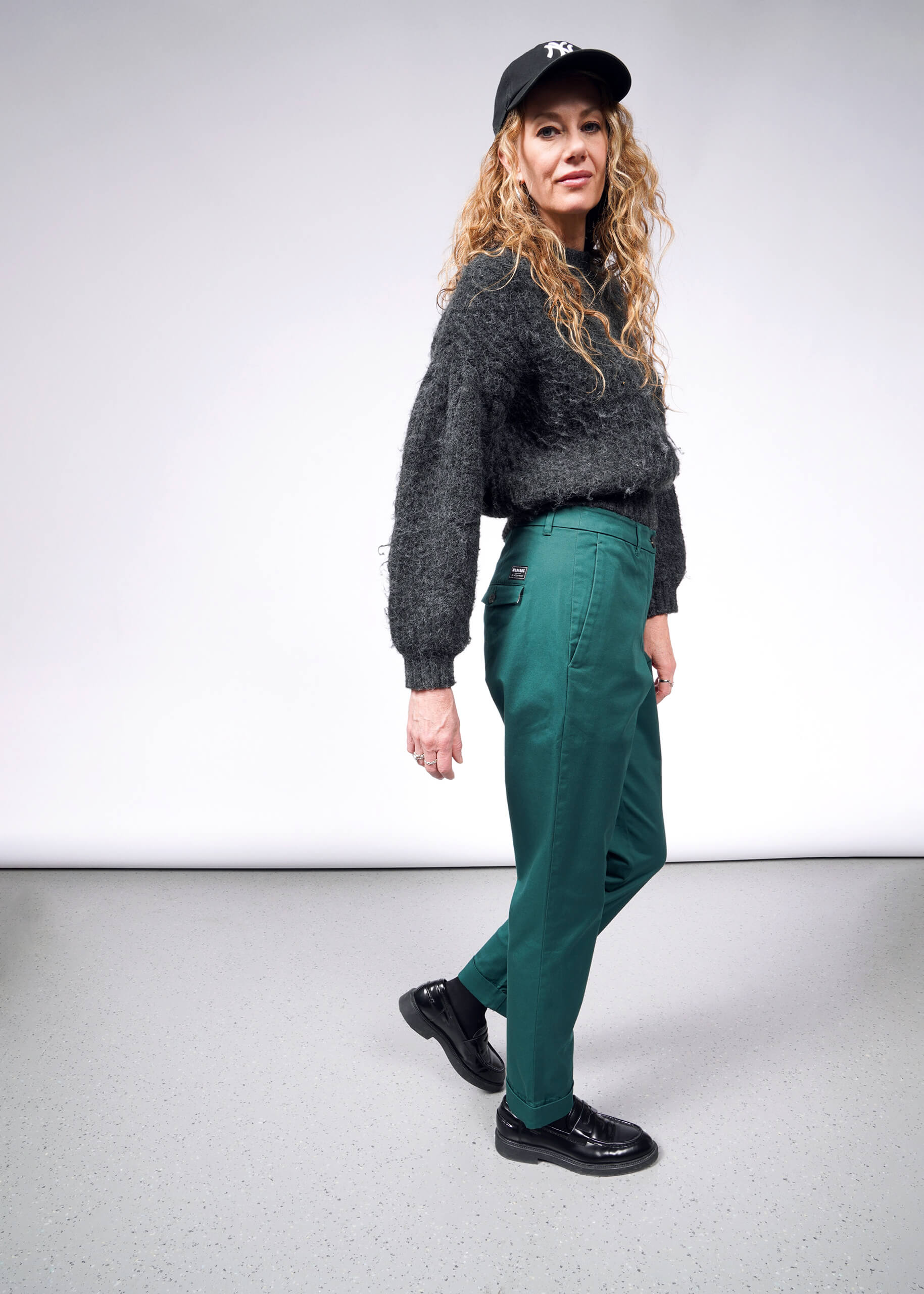 A woman with long curly hair wears a dark cap, fuzzy sweater, and The Essential Trouser in green. She poses confidently in shiny black shoes against a plain white and gray backdrop.