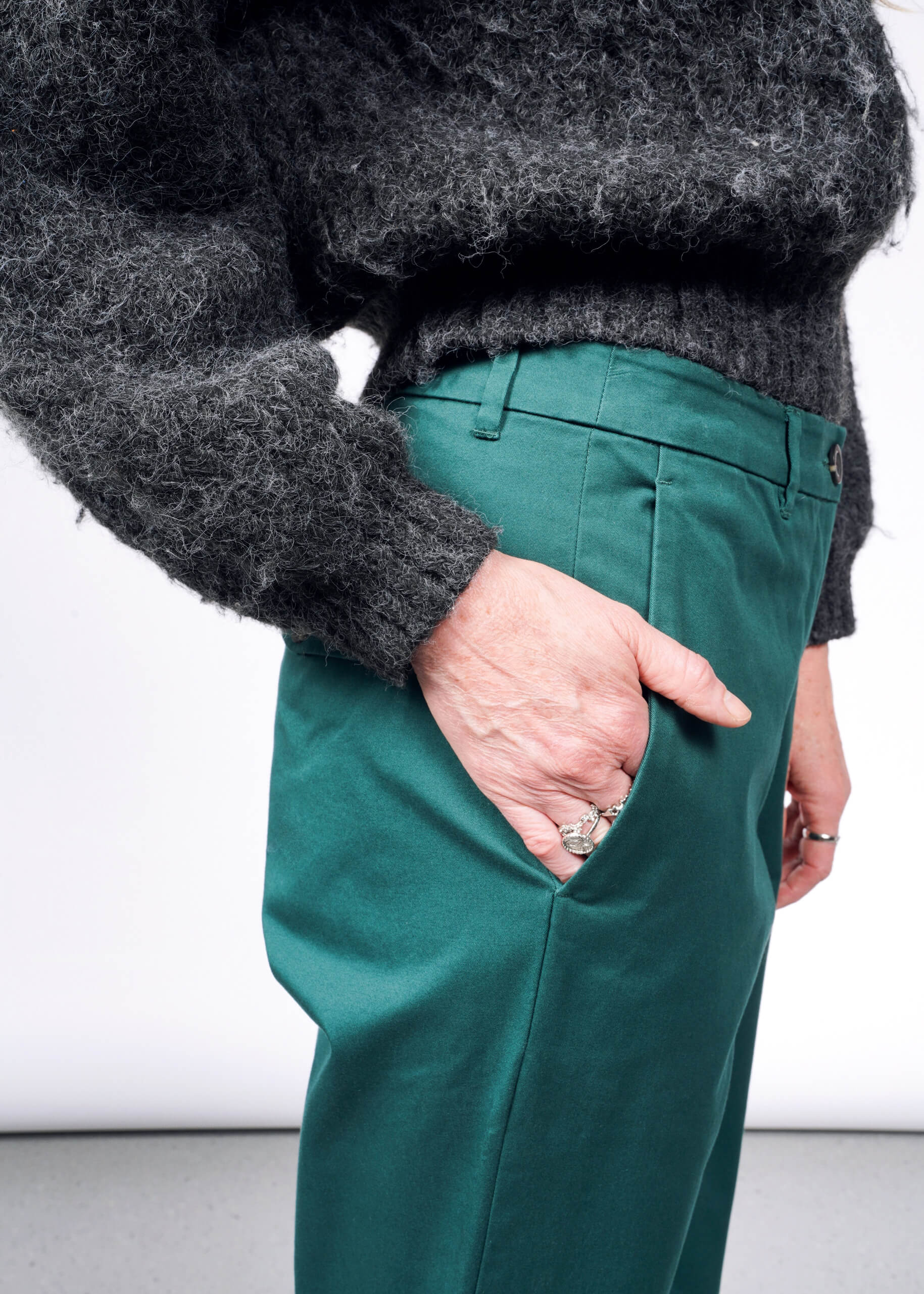 A person in a fuzzy dark gray sweater and The Essential Trouser in classic green, casually resting a silver-ringed hand in their pocket against a plain white background.