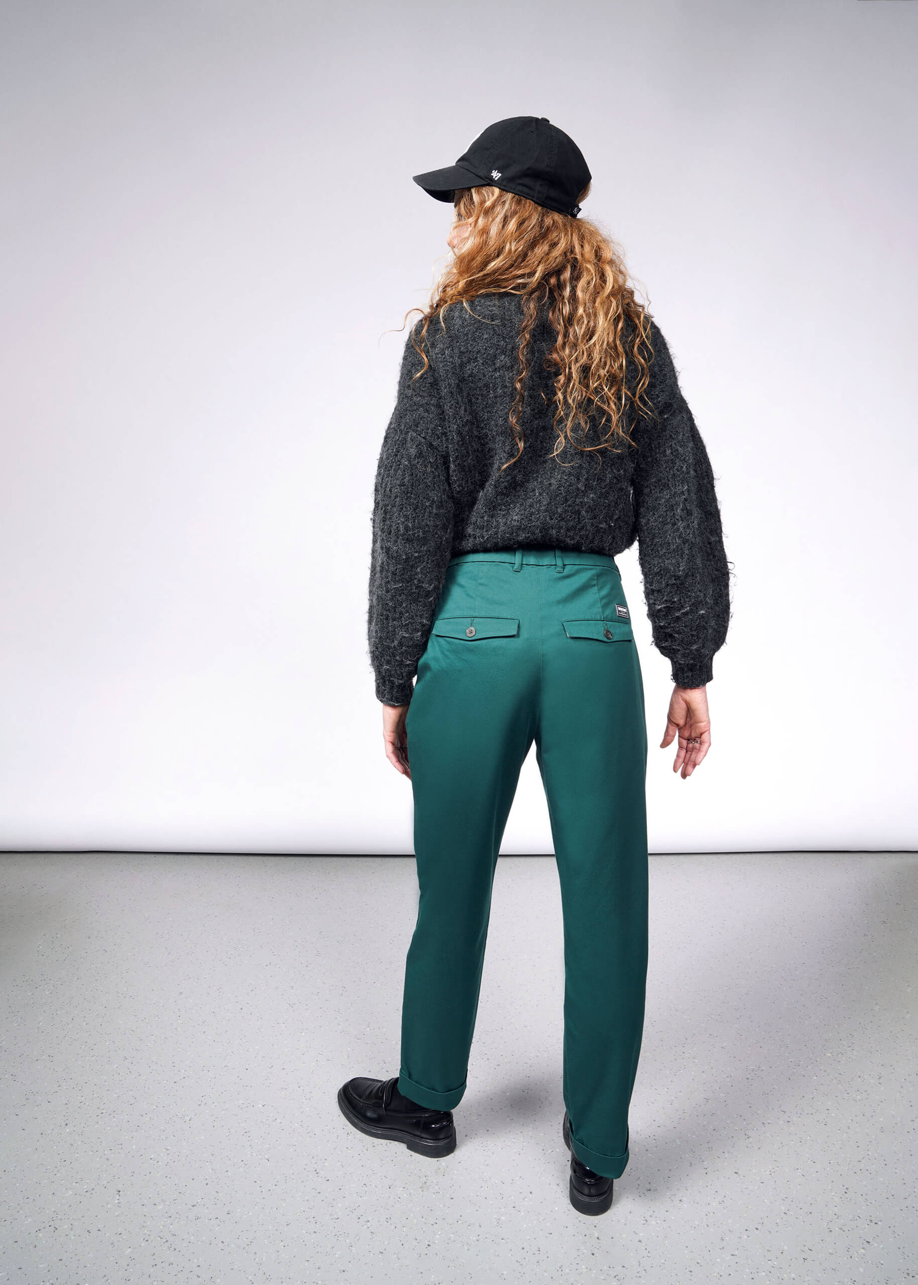 A person with long curly hair stands facing away, wearing a black cap, dark gray sweater, The Essential Trouser in green, and black shoes on a light gray floor against a plain background.