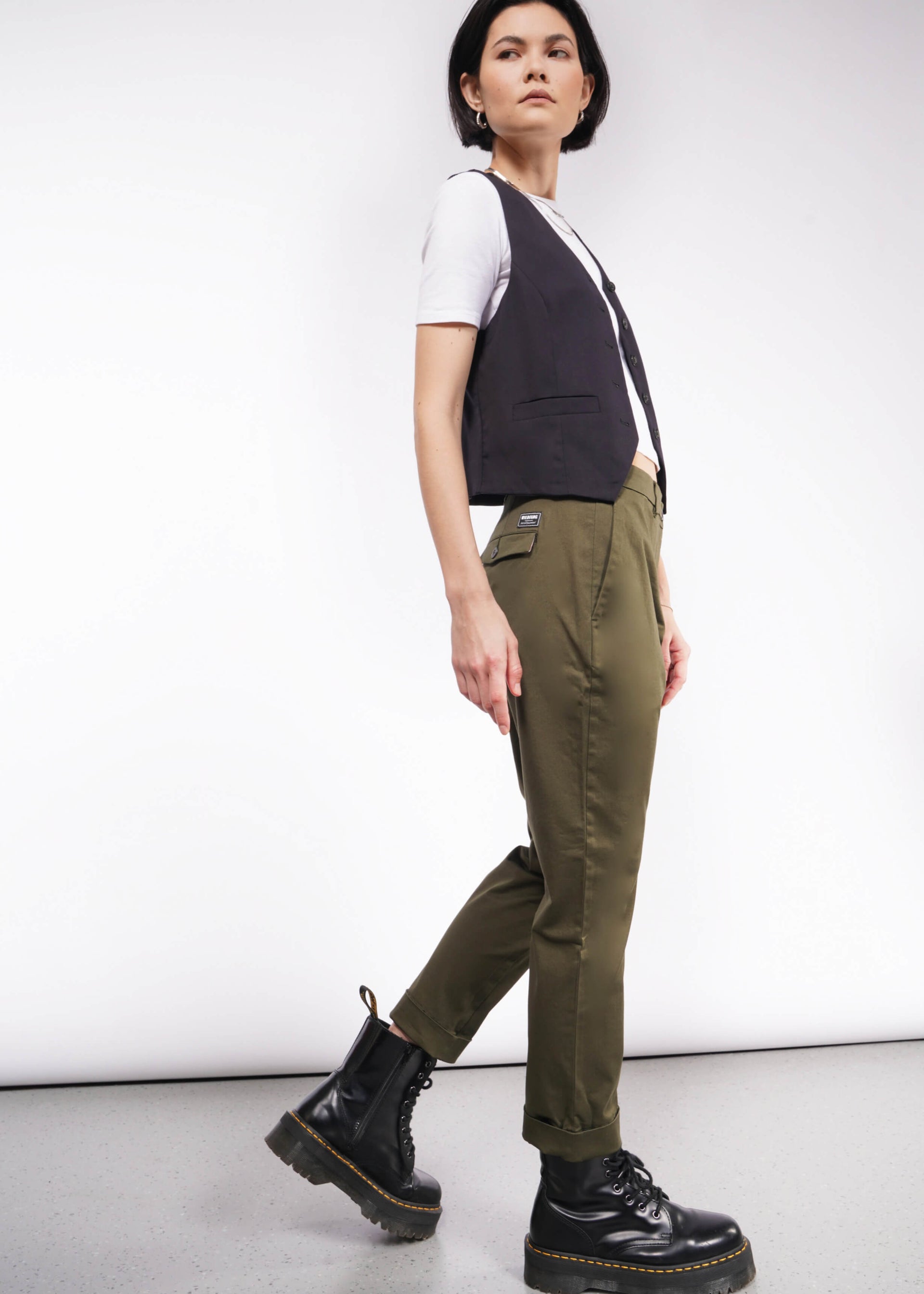 Against a plain backdrop, a person sports The Essential Trouser in olive green, paired with a white t-shirt, black vest, and black boots. They have short dark hair and glance sideways.