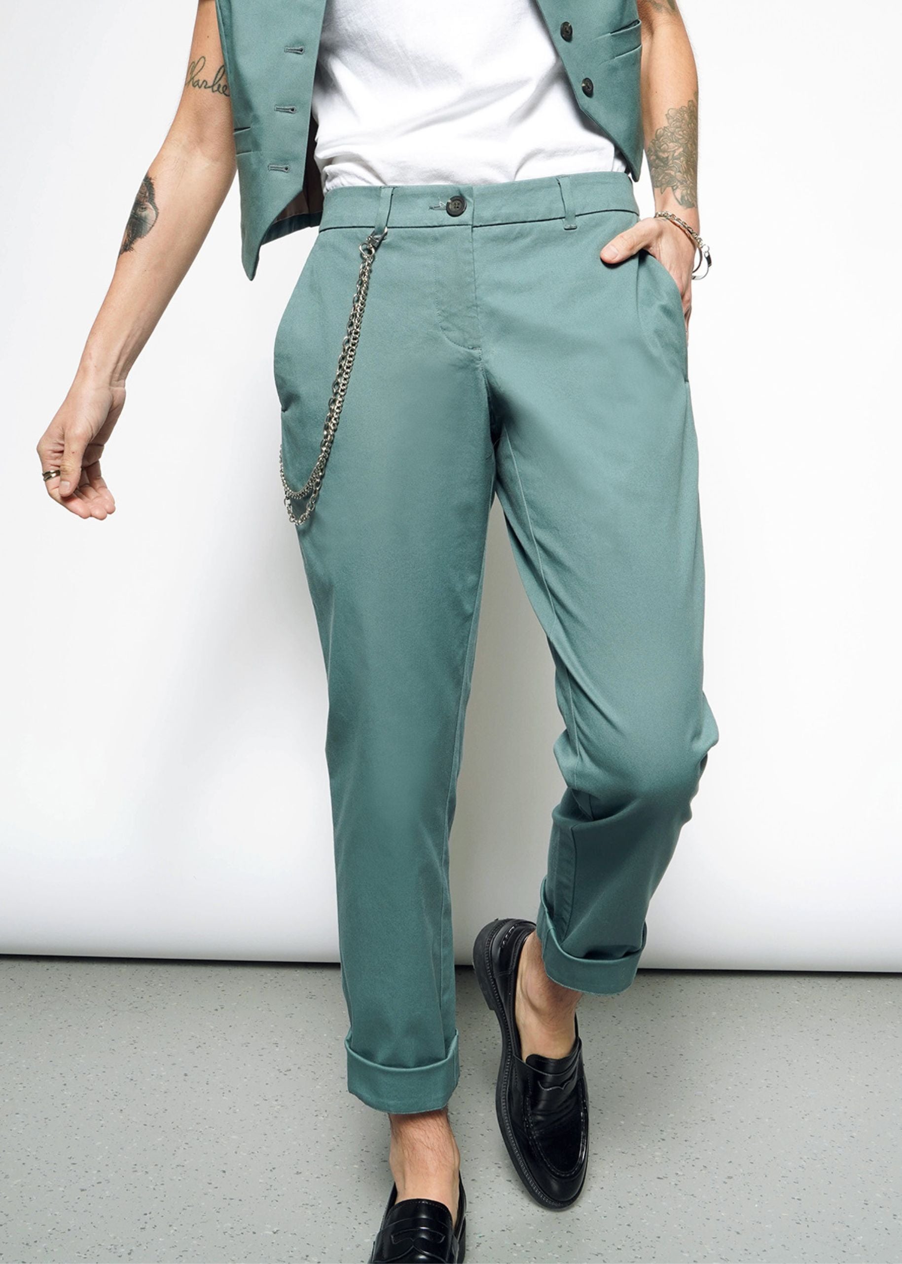 A person stands casually with a tattooed arm, hand in their pocket, wearing The Essential Trouser in classic teal with a chain accessory, paired with a white shirt and cropped teal vest. They wear black loafers with convertible cuffs against a plain white background.