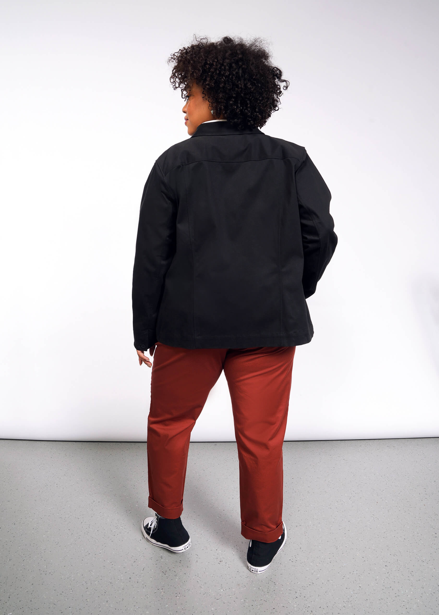 A person with curly hair, dressed in the Essential Utility Jacket with deep pockets for convenience and stretch comfort for easy movement, red pants, and black sneakers with white soles, stands against a plain white background facing away from the camera.