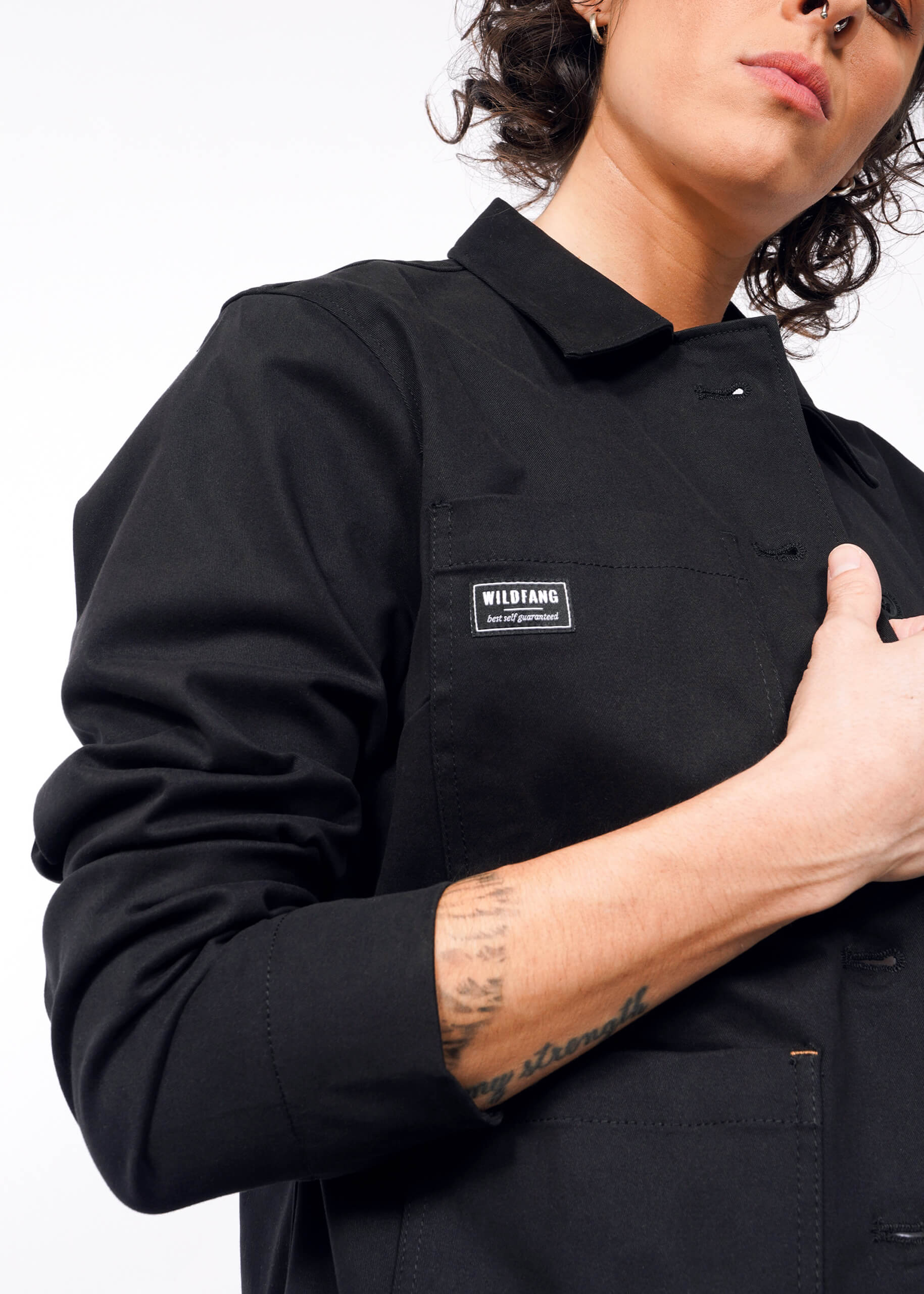 Against a plain background, a person models The Essential Utility Jacket with a Wildfang patch. The jacket features stretch comfort and deep pockets. With an arm partly raised, the intricate details of the jacket are highlighted alongside their forearm tattoo.