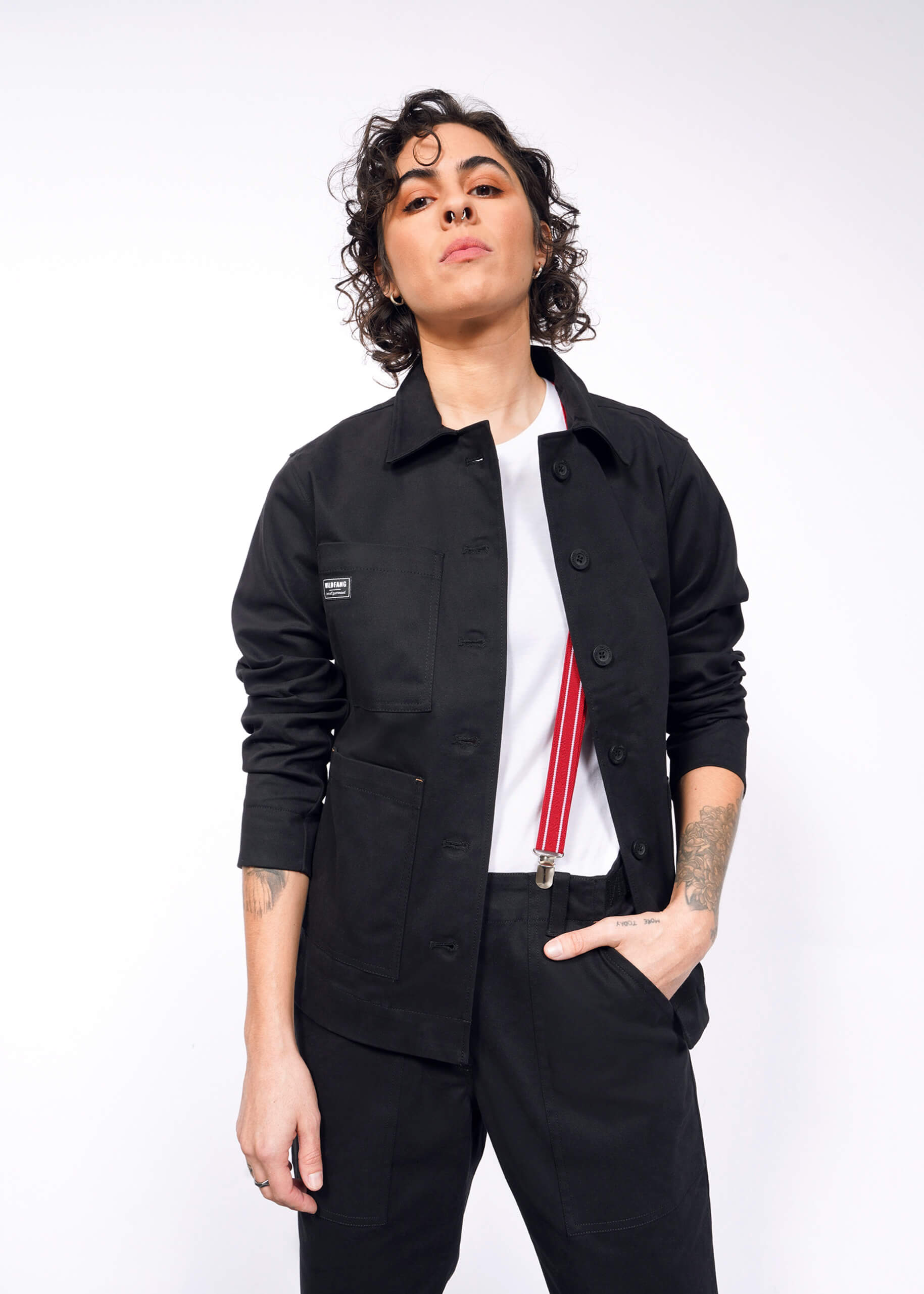 Wearing The Essential Utility Jacket and comfortable stretch pants, a person with short curly hair pairs their look with a white shirt and red suspenders, standing confidently against a white background.