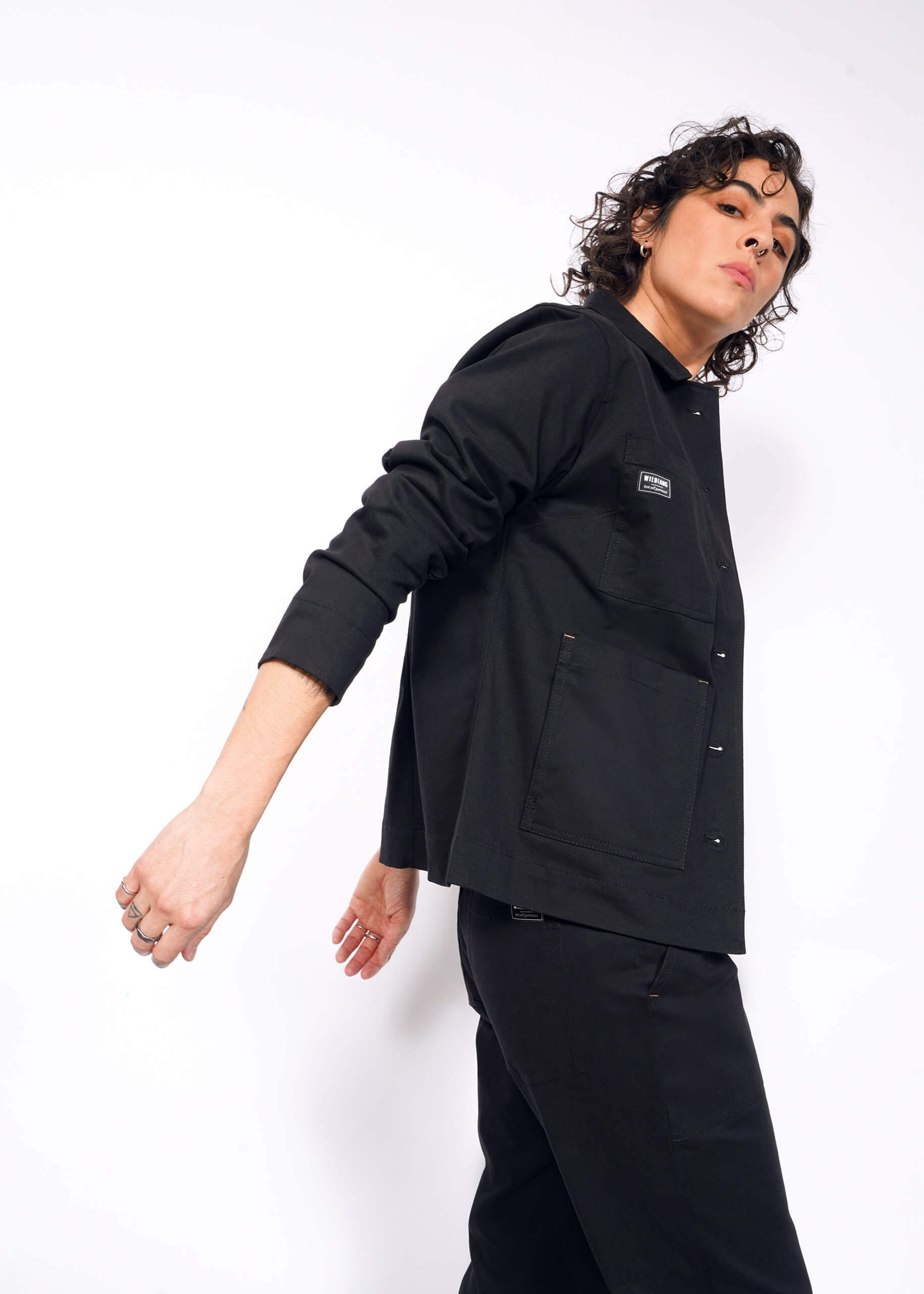 With curly hair and dressed in black, a person poses against a white background wearing The Essential Utility Jacket, featuring deep pockets and stretch comfort. Their head is slightly tilted as they gaze to the side with one arm bent.