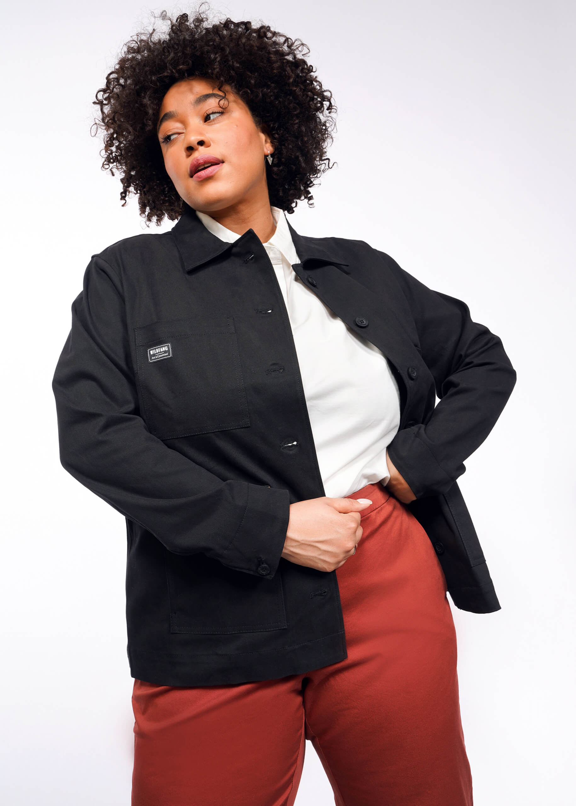 A person with curly hair models The Essential Utility Jacket, featuring deep pockets, over a white shirt and red pants. They pose with one hand on their hip, looking to the side against a plain white background.