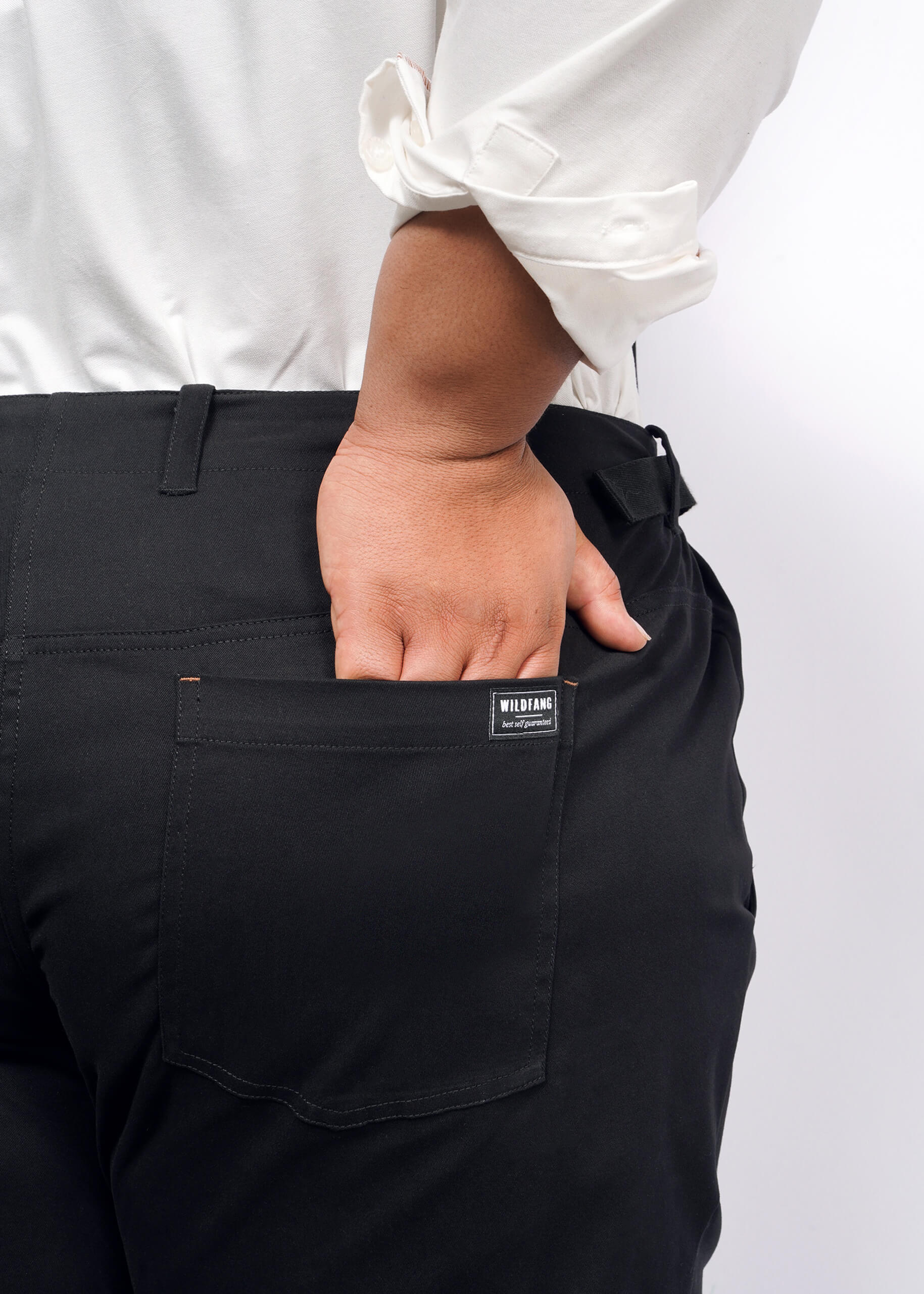 A person in a white shirt, sleeves rolled up, effortlessly embodies workwear style with their hand in the back pocket of black Essential Utility Pants. The Wildfang tag is visible against a plain backdrop, showcasing stretch cotton comfort.