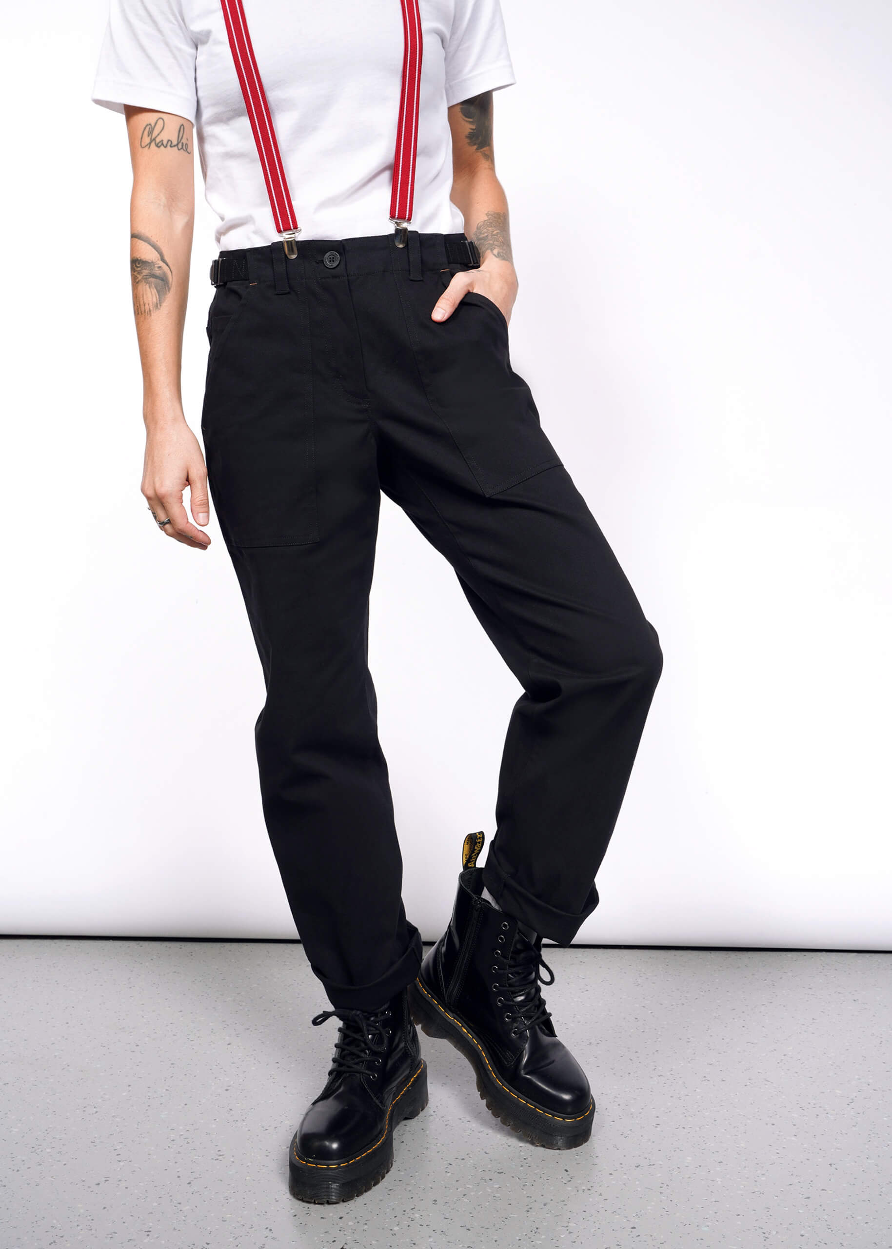 The person embodies a workwear style in black Essential Utility Pants paired with red suspenders and solid black boots. Against a plain background, their right arm rests on their hip, showcasing tattoos on both arms, while the other hand casually hangs by their side.