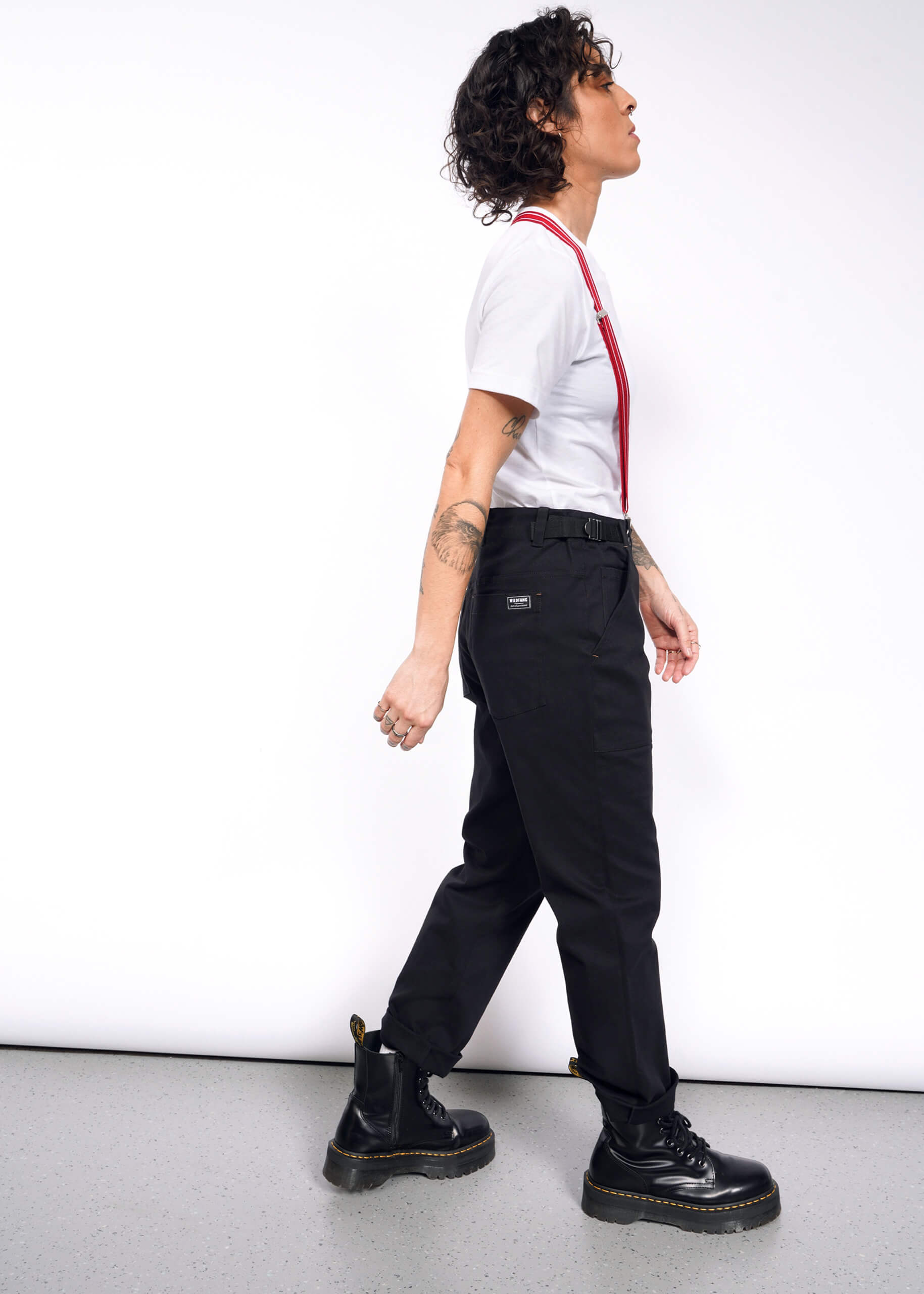 Striding confidently against a white background, a person with curly hair sports The Essential Utility Pant in black with red suspenders, paired with a white t-shirt. Their outfit blends stretch cotton comfort and workwear style, completed by black boots and tattoos visible on their arms.