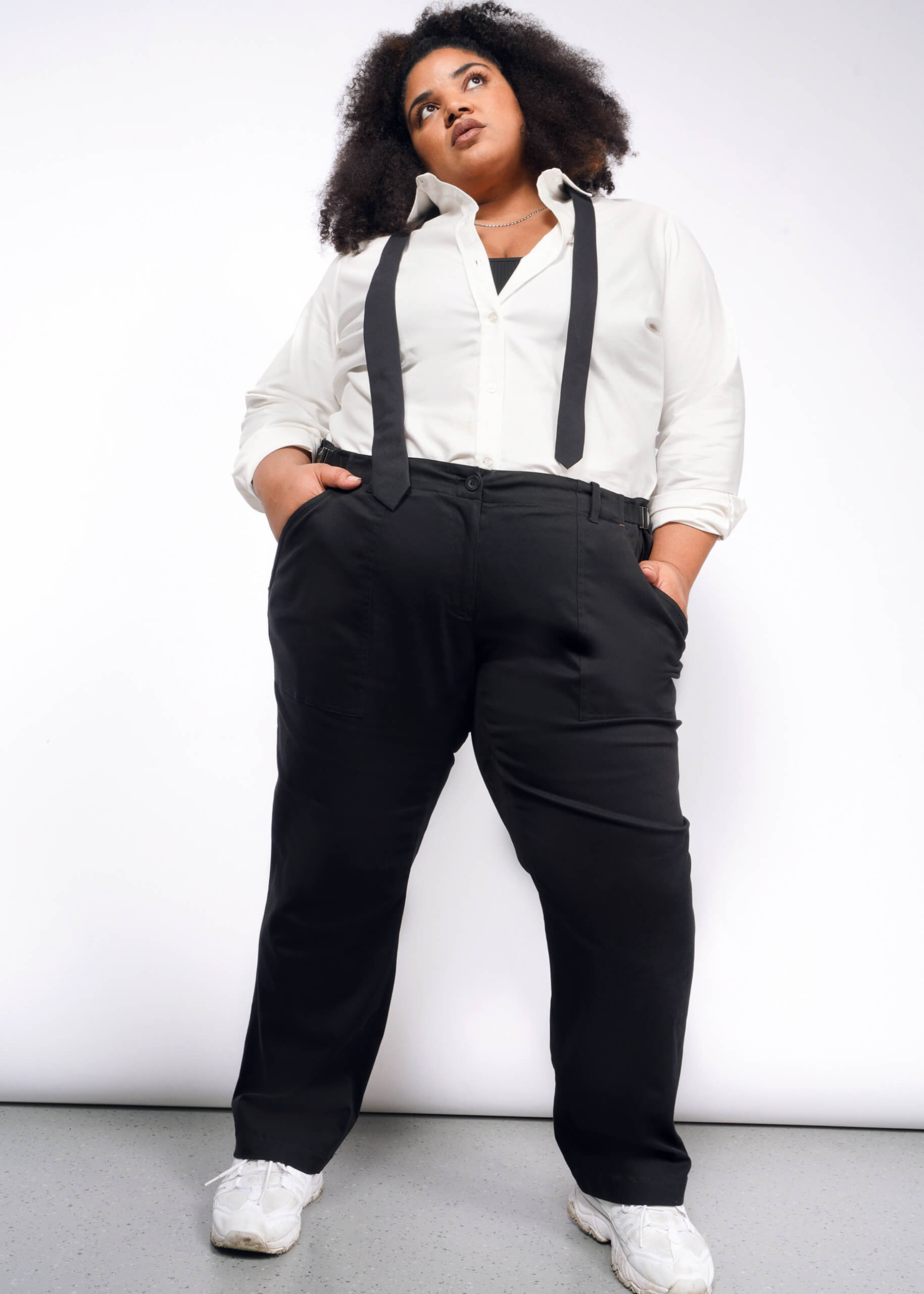 A person stands confidently against a white background, hands in pockets. Theyre wearing a white shirt, black suspenders, and The Essential Utility Pant paired with white sneakers. Their curly hair complements the workwear style as they look upwards.