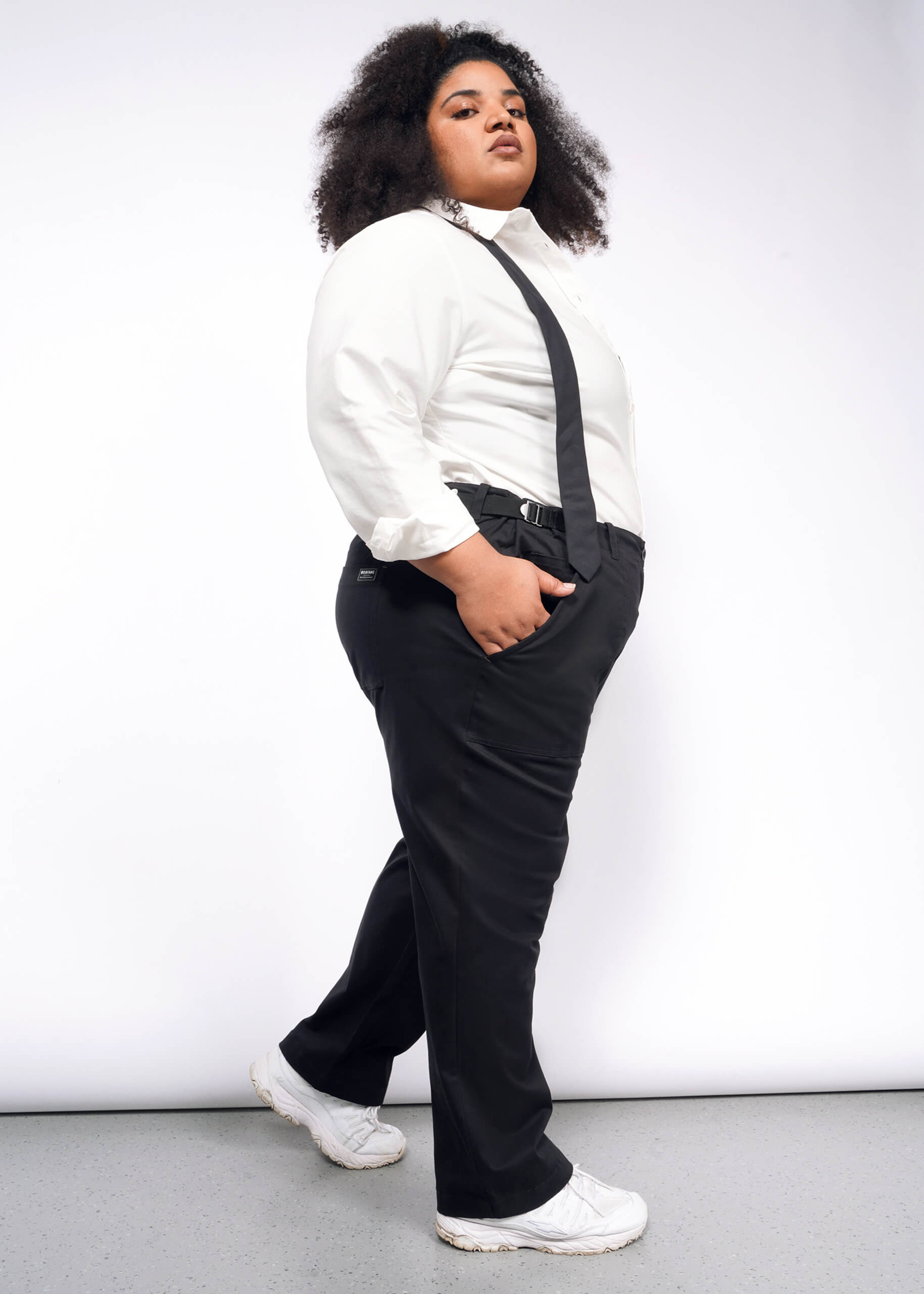 A person with curly hair in a white shirt, suspenders, The Essential Utility Pant, and white sneakers confidently poses against a plain backdrop.