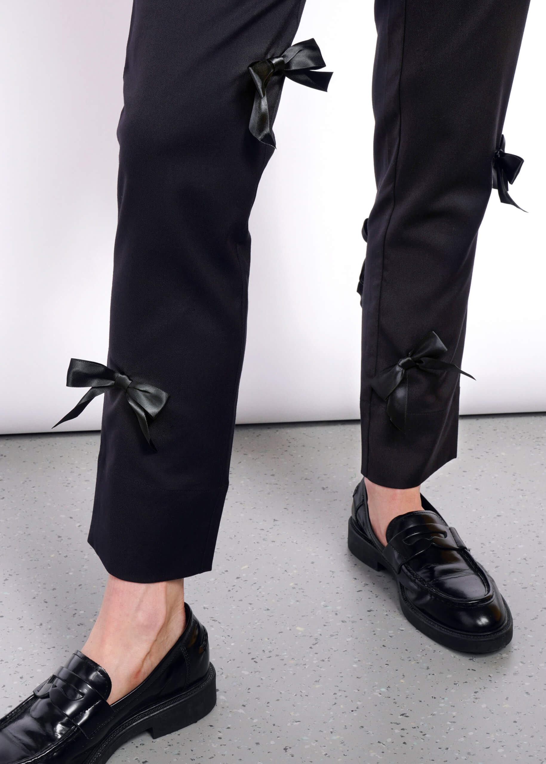 Someone wearing The Empower Bows Trouser, featuring mid-rise, black pants with exquisite bow embellishments on the sides, paired with shiny black loafers. The simple white background and speckled gray floor enhance the ultra-lux feel.