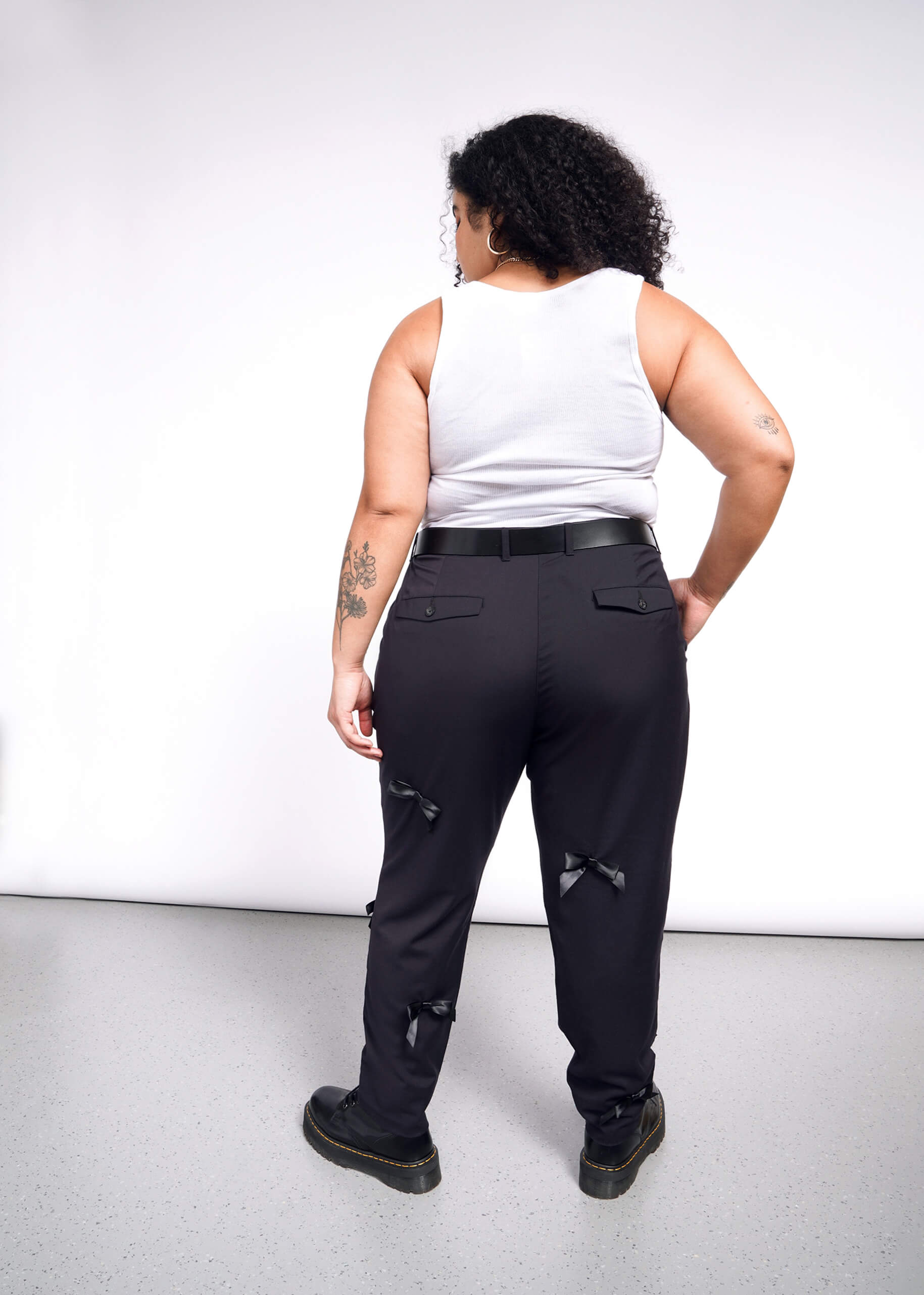 A person with curly hair stands against a white wall wearing a white tank top and The Empower Bows Trouser, ultra-luxe mid-rise black pants adorned with small bows. Tattoos cover their arms, and they complete the look with black shoes.