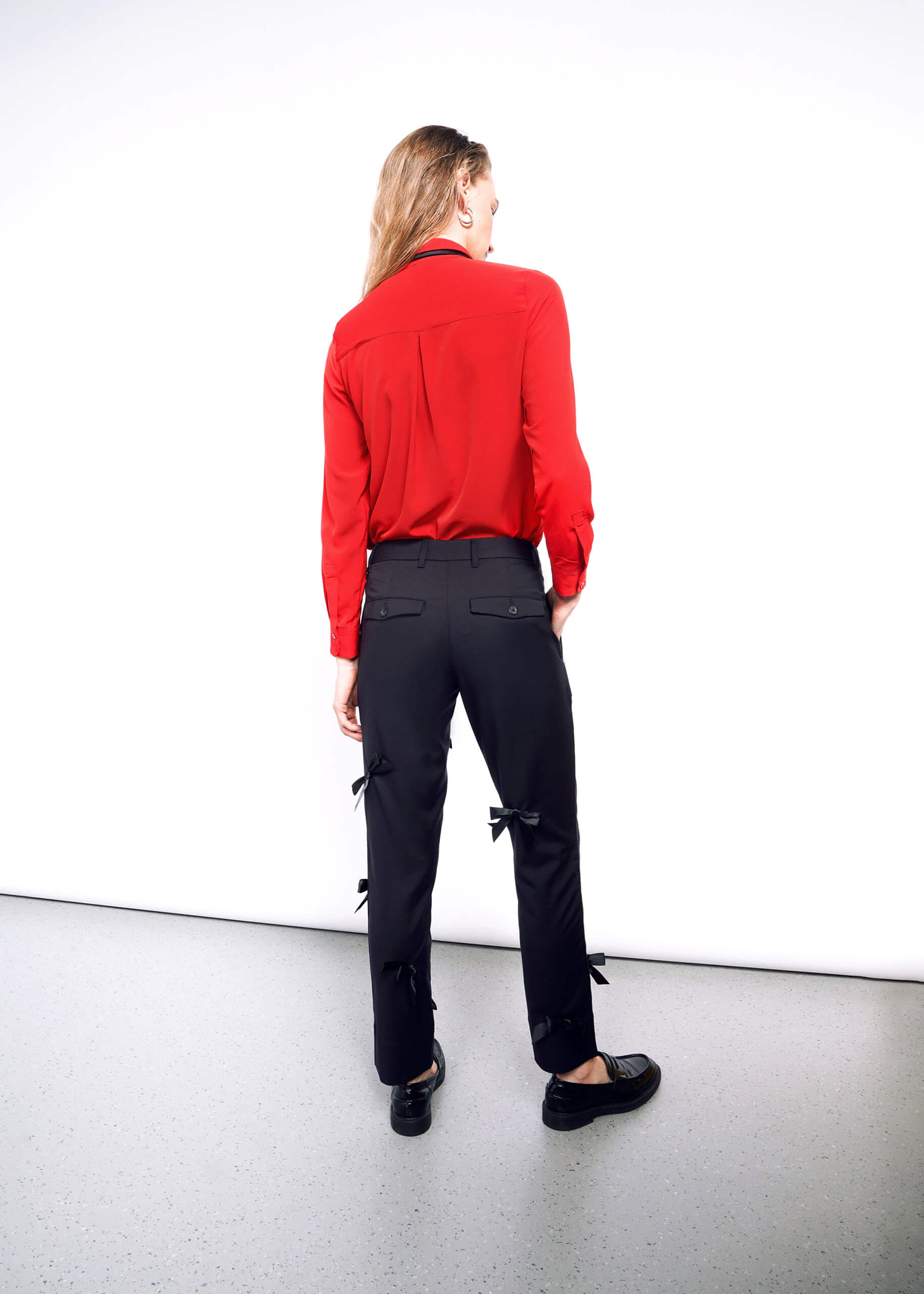 Against a white wall, a person wears an ultra-lux red long-sleeve shirt paired with The Empower Bows Trouser, featuring bow details on the sides. Facing away from the camera, they finish their stylish look with sleek black shoes.