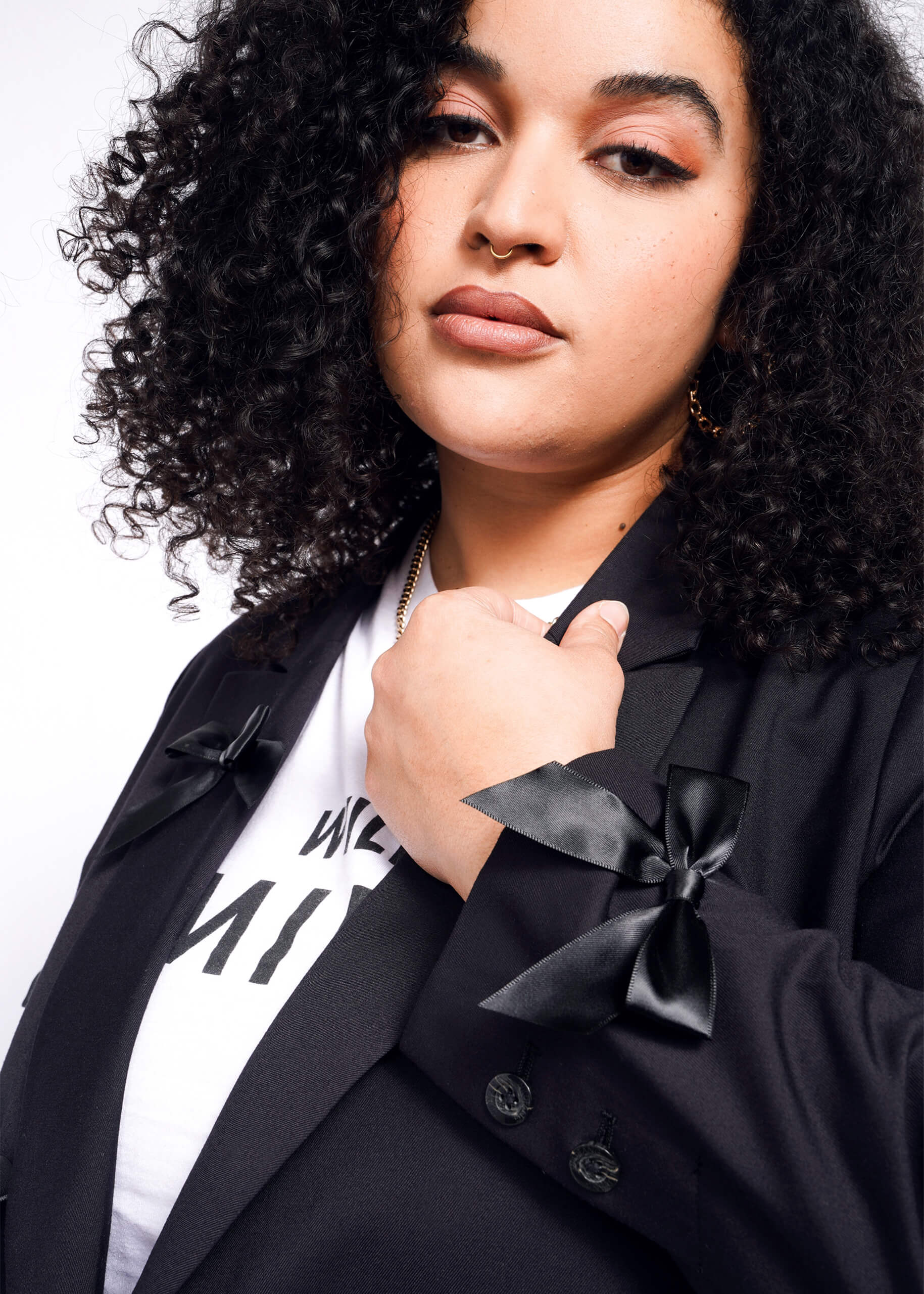 A person with curly hair poses confidently in The Empower Bows Double Breasted Blazer, featuring tailored oversized design and bow details on the sleeves, over a white T-shirt against a white background.