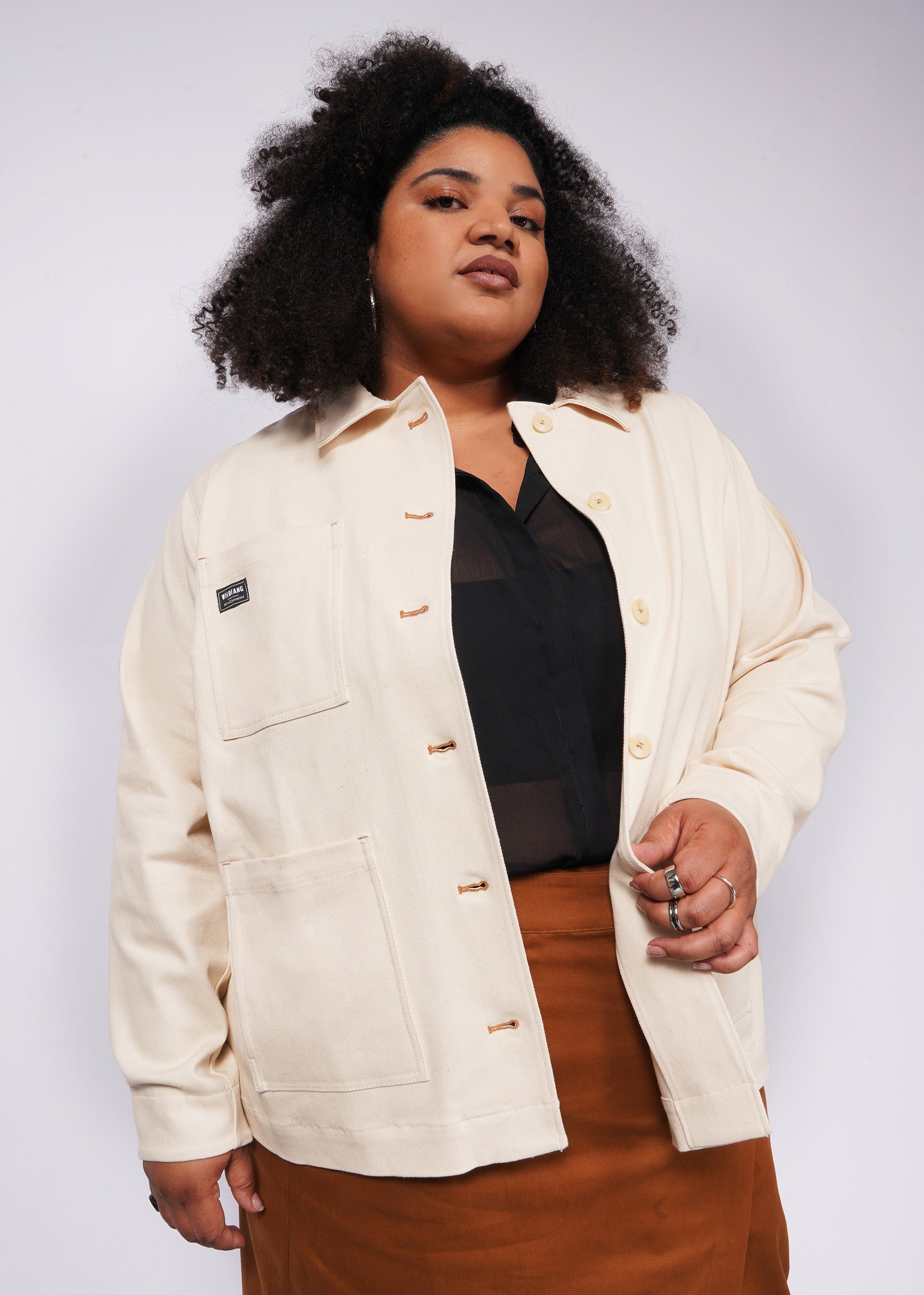 A person with curly hair poses confidently, wearing The Essential Denim Utility Jacket over a black top and a brown skirt. The cream-colored jacket features deep pockets and a label, set against a plain, light background.